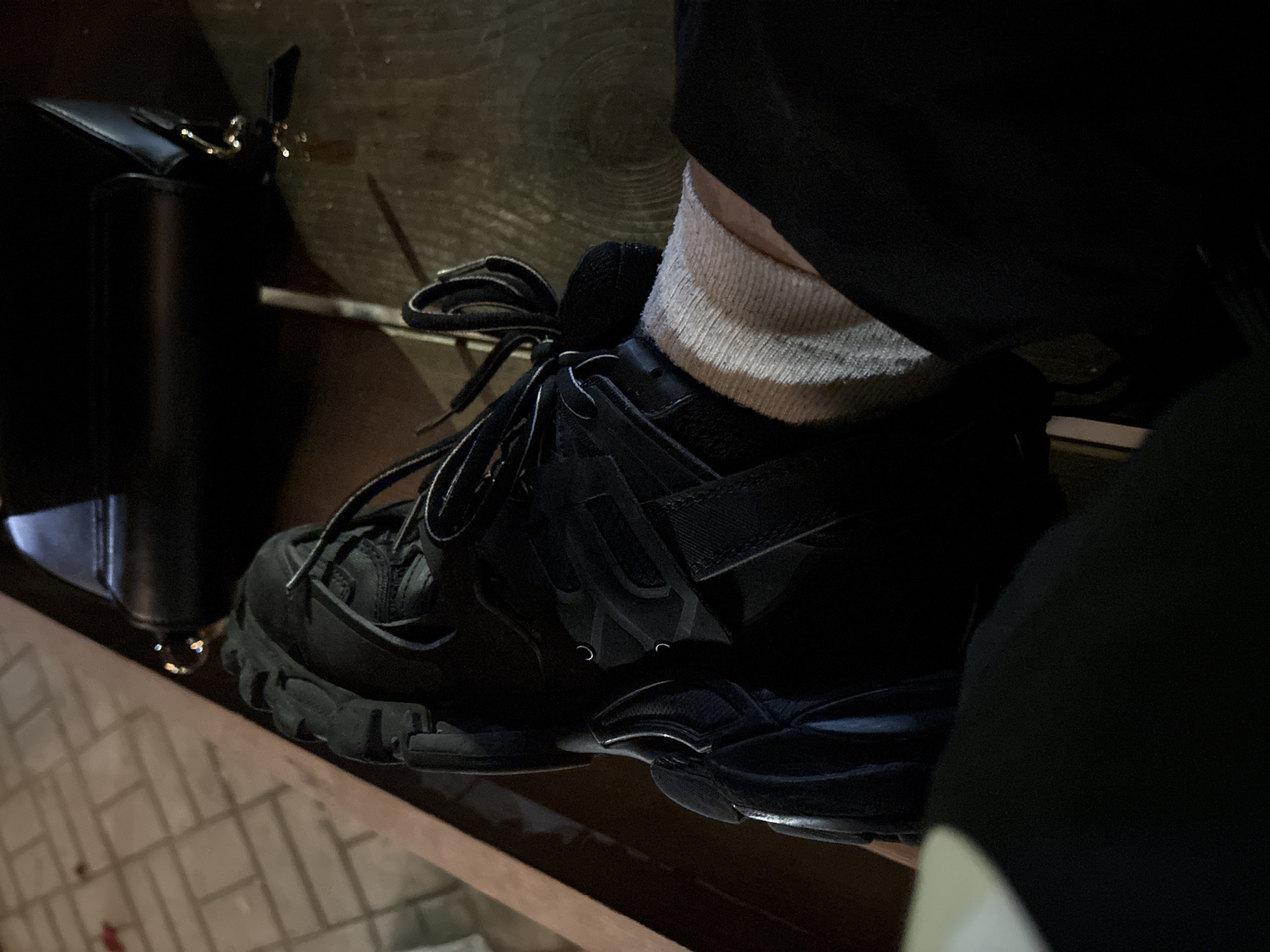 A close-up shot captures a dark athletic shoe on a wooden surface, likely indoors. The shoe is primarily black, with visible laces and a thick sole. A white sock is visible above the shoe's opening. To the left of the shoe, a black purse is positioned. The background is blurry, suggesting a shallow depth of field. The overall lighting is subdued.