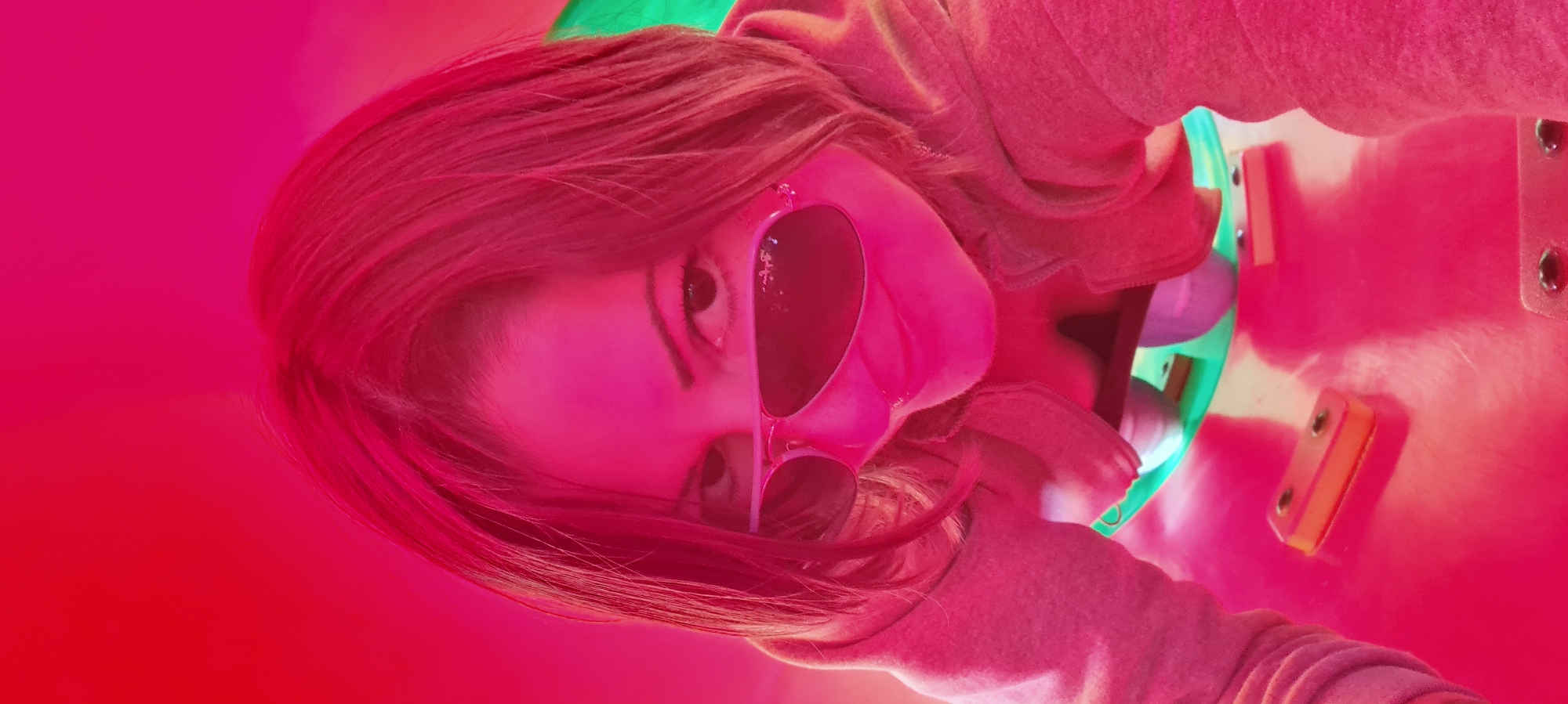 A person with reddish-brown hair is visible, wearing heart-shaped sunglasses. They appear to be inside a playground tunnel or slide, indicated by the pink and green plastic surfaces surrounding them. The lighting is predominantly red. The scene is likely outdoors. No identifiable landmarks or other details confirm the location as Lachish Regional Council, Israel.