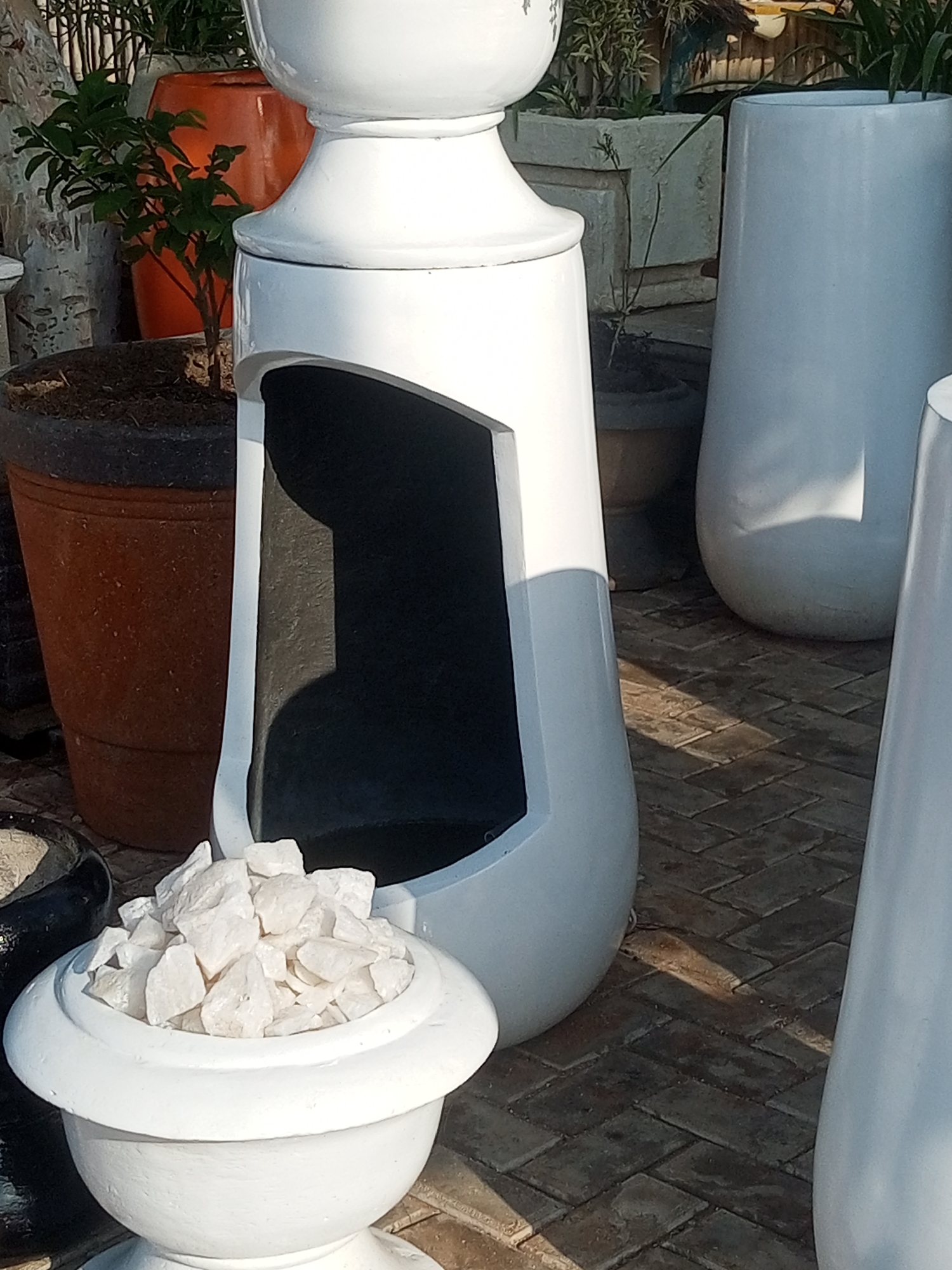 In Gombe, Nigeria, an outdoor scene showcases a collection of white ceramic planters and decorative items, bathed in the warm light of what appears to be late afternoon or early morning. The primary subject is a large, hollow white ceramic planter with a unique cut-out opening, revealing a dark interior. Next to it, a smaller white planter is filled with rough, white stones, suggesting a decorative arrangement.
In the background, other planters are visible, including a tall, light blue cylinder and a large orange pot. A small green plant with dark leaves is situated in a dark brown pot, adding a touch of natural color to the otherwise monochromatic ceramic display. The planters are arranged on a brick-paved surface, which is partially covered in sunlight and shadow, indicating clear weather.
The overall impression is one of a retail or display area for garden or home decor, focusing on ceramic pots and decorative stones. There are no people visible in the image, and no text is present. The scene is static, with no activity taking place. The lighting suggests a pleasant day, likely outdoors due to the brick paving and the presence of plants.