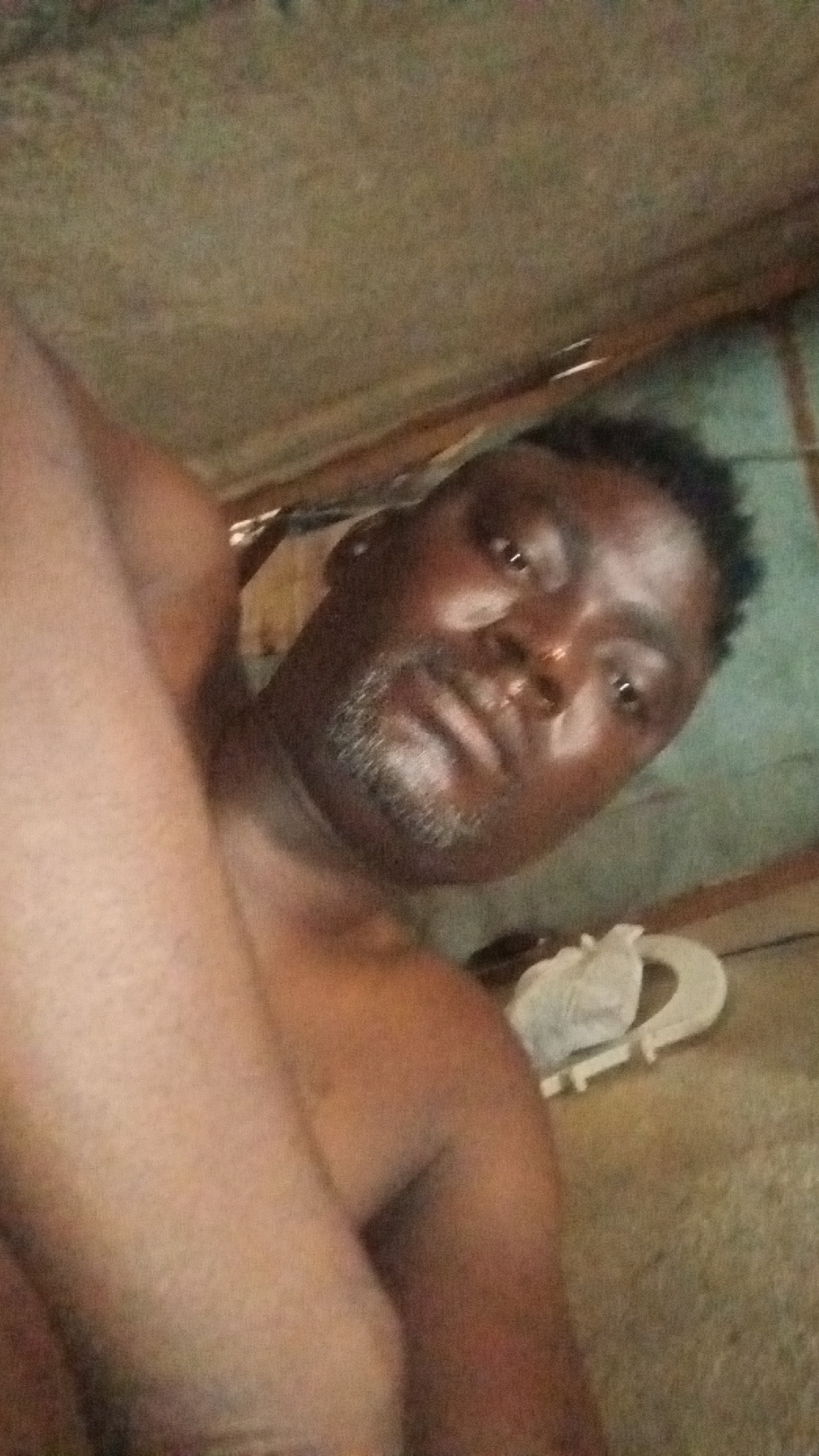 The image shows a dark-skinned man lying down. He has short black hair and a patchy beard. His skin appears shiny with possible perspiration. He is topless, and part of his arm is visible in the lower left corner of the frame. Behind the man, there are a pair of white sandals. The background suggests a simple interior space with possibly wooden or metal walls and a rough ceiling. The location is listed as Unknown, Unknown.