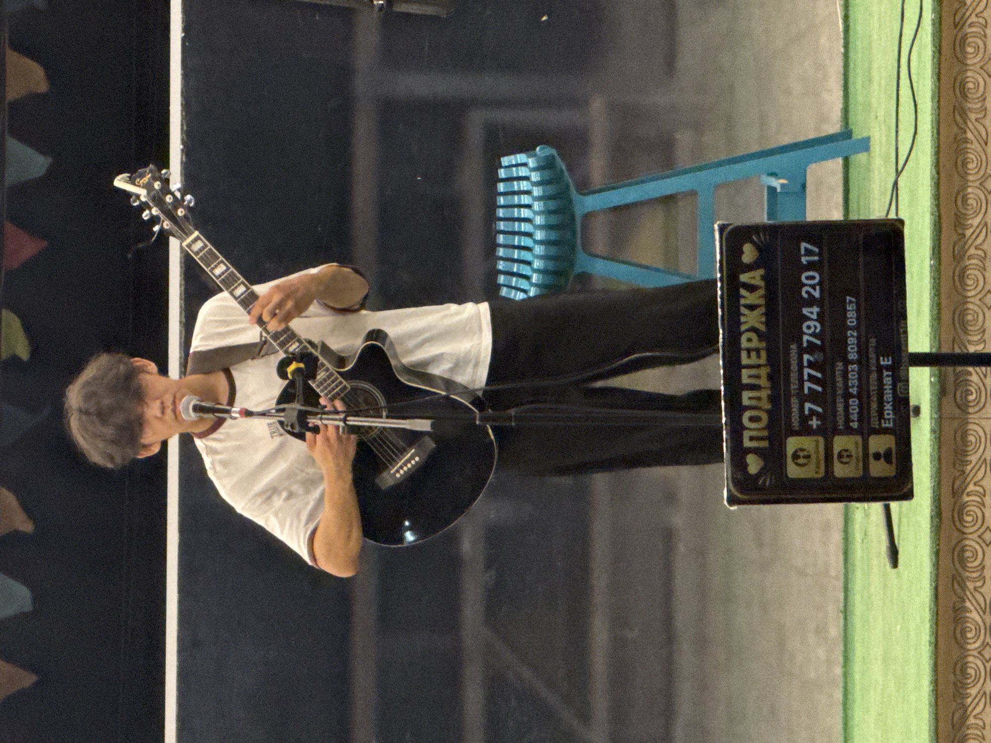 A young male musician stands on a stage, playing an acoustic guitar and singing into a microphone. He is wearing a white t-shirt with a maroon collar and dark pants. Behind him, a bright blue stool is visible. In the foreground, a dark sign with yellow lettering displays contact information, including a phone number and card details, for "PODDERZHKA" (support). The background appears to be a dark wall adorned with colorful triangular flags. The overall setting suggests an outdoor event or performance in Pavlodar, Kazakhstan, as indicated by the Cyrillic script on the sign.