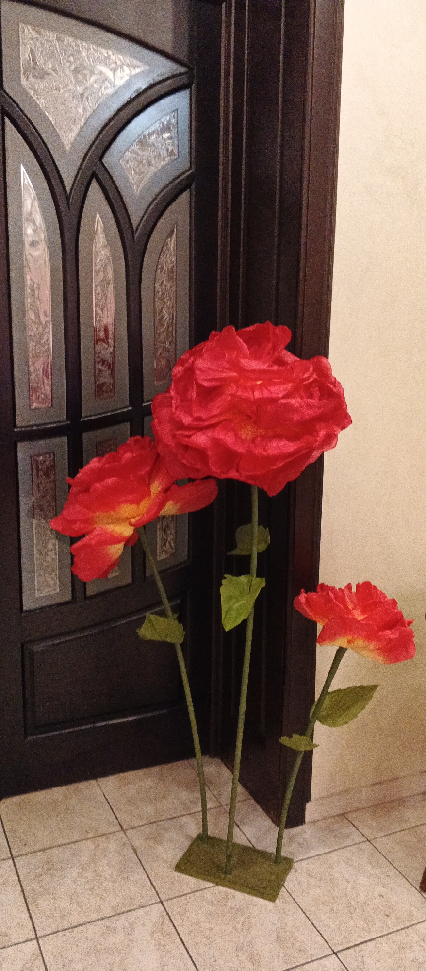 Three large, artificial red flowers with yellow centers stand on a green base in front of a dark brown door with decorative glass panels. The flowers are positioned at different heights and angles, with one large bloom at the top, a medium-sized bloom to the left, and a smaller bloom to the right. The door is ornately designed with geometric patterns and frosted glass inserts. The floor is tiled with light-colored square tiles. The overall scene suggests an indoor setting, possibly a home or a public building, in Mukachevo, Ukraine. There are no people visible in the image.