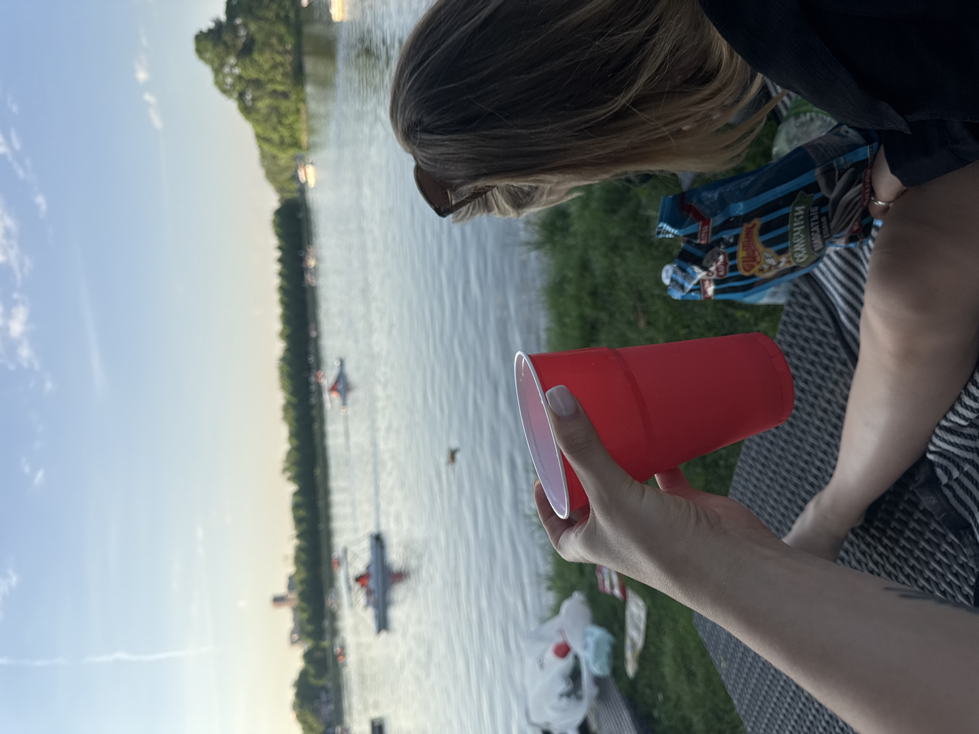 A sideways, eye-level shot shows an outdoor scene at a waterfront location in Moscow, Russia. In the foreground, a person holds a red plastic cup filled with a reddish liquid. A bag of sunflower seeds with Russian text is near the person. Further away, a woman with blonde hair and sunglasses is visible. A body of water fills the background, with several rowing boats visible in the distance. The banks of the river are lined with trees. The sky is light, suggesting it is either early morning or late afternoon.