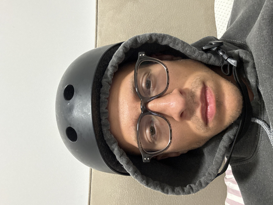 A male individual is depicted in a close-up, head-and-shoulders portrait, facing directly towards the camera. He wears a dark grey hooded sweatshirt with the hood pulled over his head. A black sports helmet, exhibiting minor surface scuffs and two circular ventilation holes on its front, is worn over the hood. He wears eyeglasses with dark, patterned frames. Short stubble is visible on his chin and upper lip. His eyes are open, and his mouth is closed. A plastic cord lock is visible on the drawstring of his hoodie on the right side of the frame. The background consists of a plain, light-colored wall in the upper portion, and sections of light-colored upholstered furniture (likely a sofa or headboard) flanking the subject. A small portion of striped fabric, possibly bedding, is visible at the bottom left. The scene is set indoors within a residential-type environment. No specific geographical markers are present to identify the location as Tel-Aviv, Israel.
