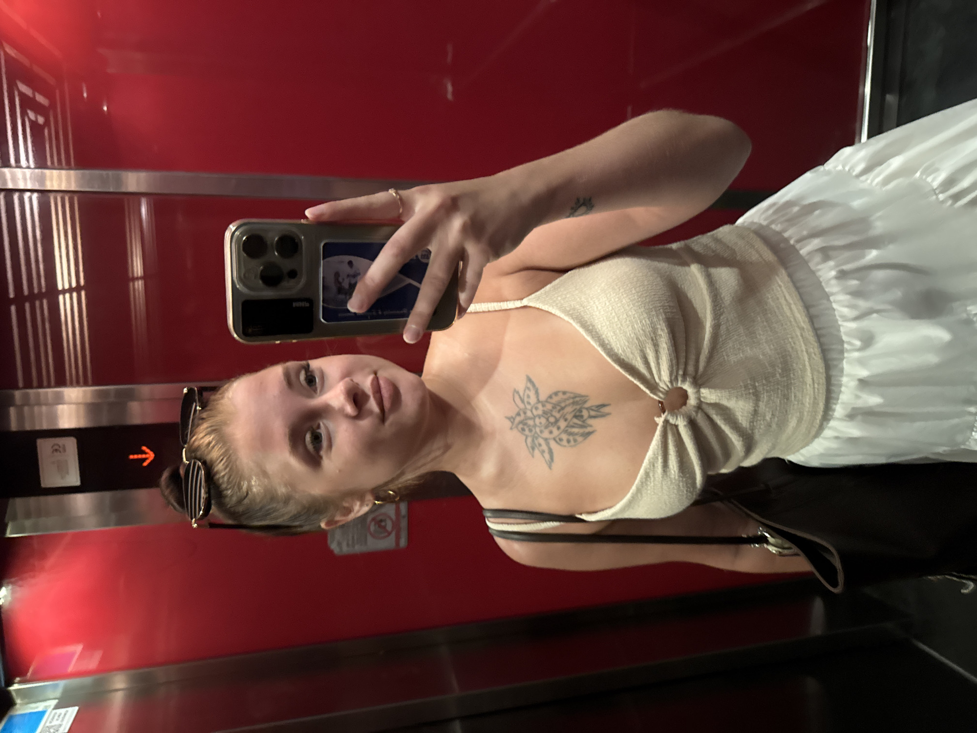 A young woman with her hair pulled up in a bun and sunglasses on her head takes a selfie in an elevator. She is wearing a light-colored, sleeveless top with a knotted front and a white, ruffled skirt. A dark-colored bag is slung over her left shoulder. She has a tattoo on her chest and a ring on her right ring finger. The elevator has red, reflective walls and a brushed metal panel. The woman is looking at her phone, which is held in her right hand. There are signs on the elevator wall, one of which is an orange arrow pointing down. The location is identified as Aksu, Turkey.