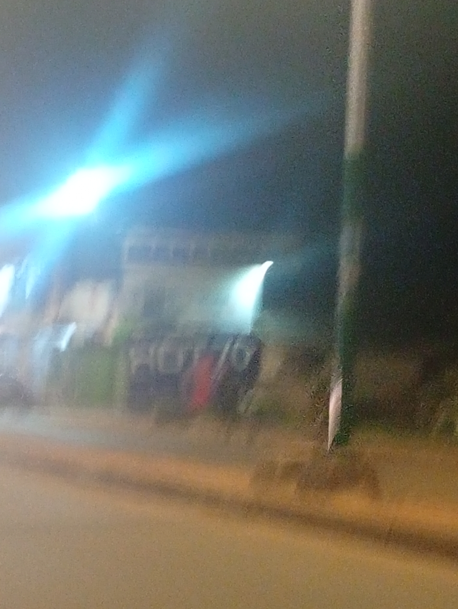 The image is a blurry, night-time photograph taken from a moving vehicle, likely on a street in Bukuru, Nigeria. The primary focus is a bright, bluish-white light source emanating from the left side of the frame, casting a strong glare across the scene. In the mid-ground, there appears to be a building with visible windows, partially obscured by the light. In front of the building, several blurry objects that might be vehicles or structures are visible, with some hints of color such as red and green. A vertical pole, possibly a streetlight or signpost, is prominent on the right side of the image. The foreground consists of a blurred road surface, suggesting the photograph was taken while the vehicle was in motion. The overall impression is one of a somewhat desolate urban environment at night, with artificial lighting playing a significant role in the composition.