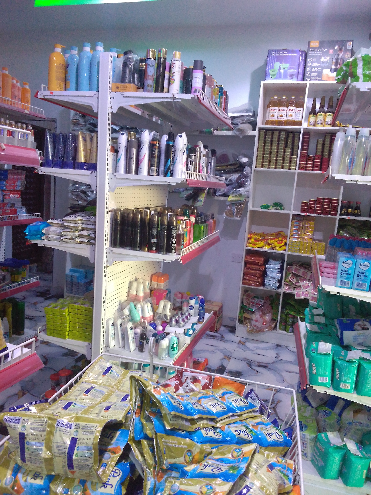The image depicts the interior of a small retail store, likely a pharmacy or convenience store, located in Howayi, Nigeria. The shelves are stocked with a variety of products, including personal care items, cleaning supplies, and food products.
On the left side of the image, shelves are filled with bottles of shampoo, lotion, and other toiletries. There are also packaged goods such as soap and toothpaste. The middle section of the store features shelves stocked with spray cans, likely deodorants or air fresheners, and various packaged snacks and food items. The right side of the image shows a multi-compartment shelving unit filled with bottled beverages, canned goods, and boxes of feminine hygiene products. In the foreground, there are several large baskets overflowing with packaged food items, predominantly what appear to be powdered milk sachets and other dry goods.
The flooring is tiled with a marble-like pattern. The lighting appears to be overhead fluorescent, casting a bright, slightly cool tone over the scene. There are no people visible in the image. The overall impression is one of a well-stocked, albeit somewhat crowded, retail environment.