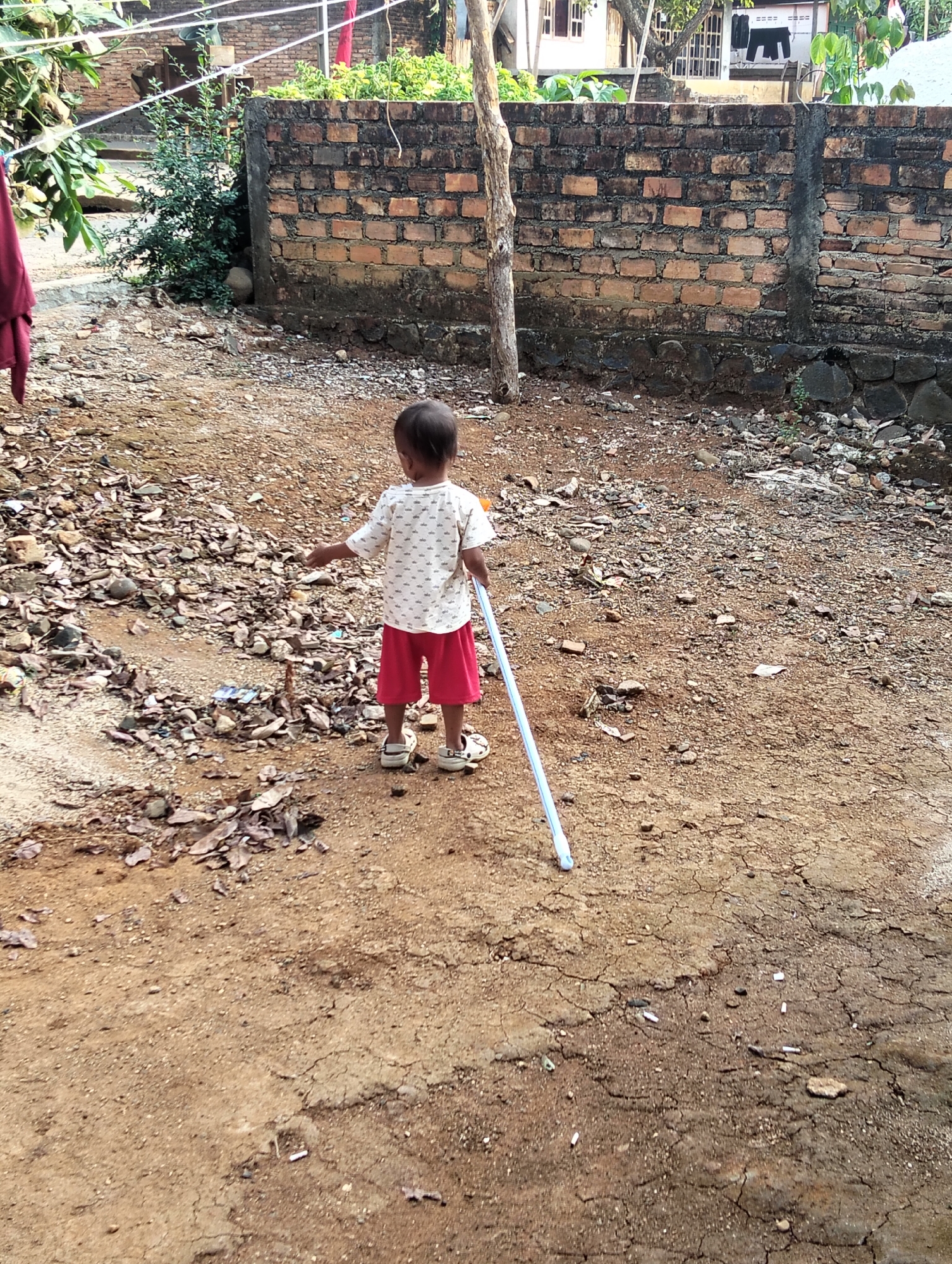 A young child, viewed from behind, stands on a dirt path in a rural or undeveloped area. The child is wearing a white t-shirt with a printed pattern and red shorts, along with sandals. They are holding a light blue pole with a white tip. The ground is a mix of dirt, small stones, and scattered dry leaves. In the background, a brick wall runs horizontally, with vegetation growing behind it and a tree trunk in the foreground. The location is identified as Tanjung Harta, Indonesia.