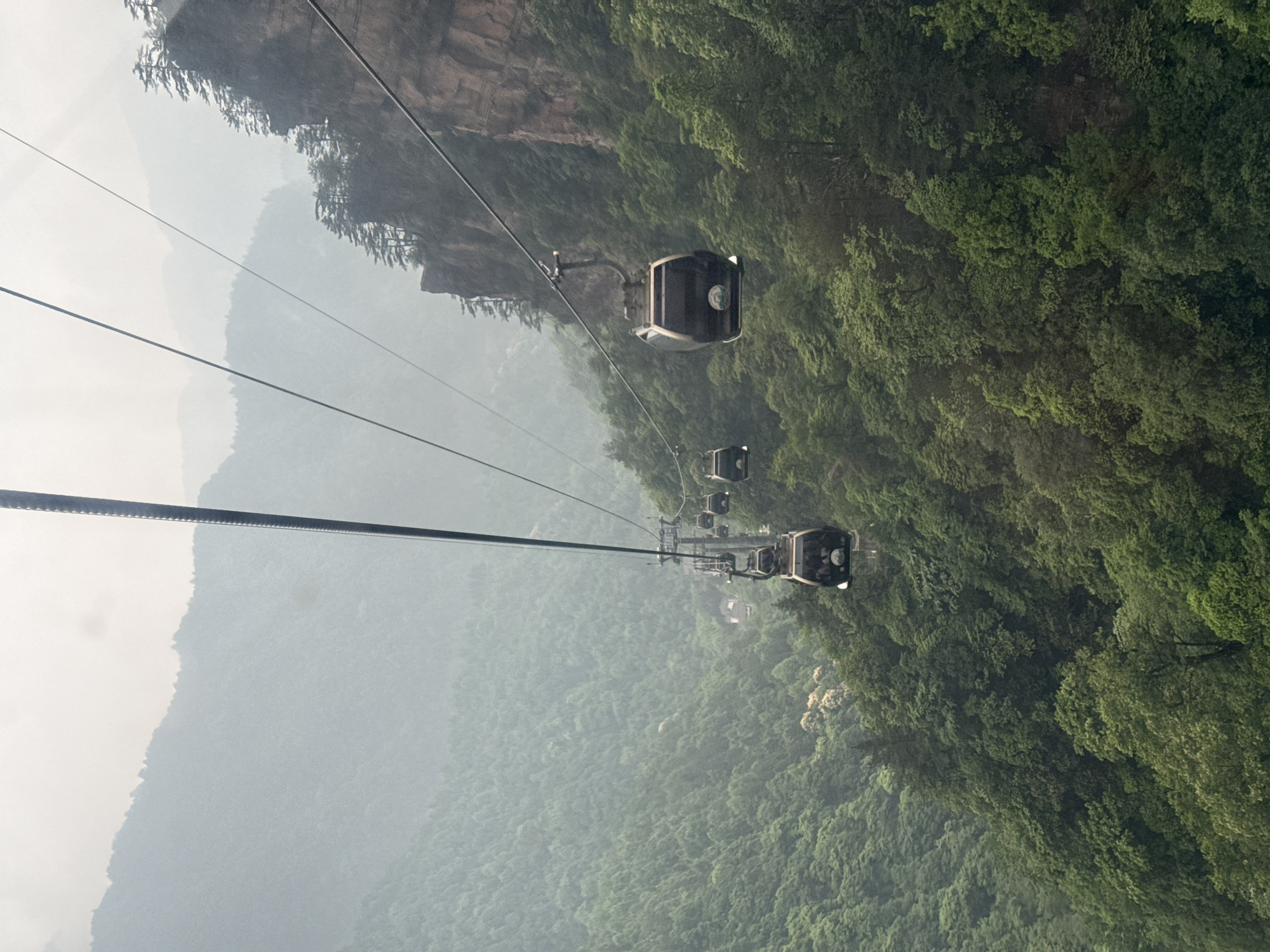 The image shows a series of cable cars traveling over a dense, green mountainous terrain. The scene is hazy, with fog obscuring the distant landscape. The cable cars are the main subject, with a clear view of the cables that support them. The location is Wulingyuan, China, as determined by the distinctive landscape.