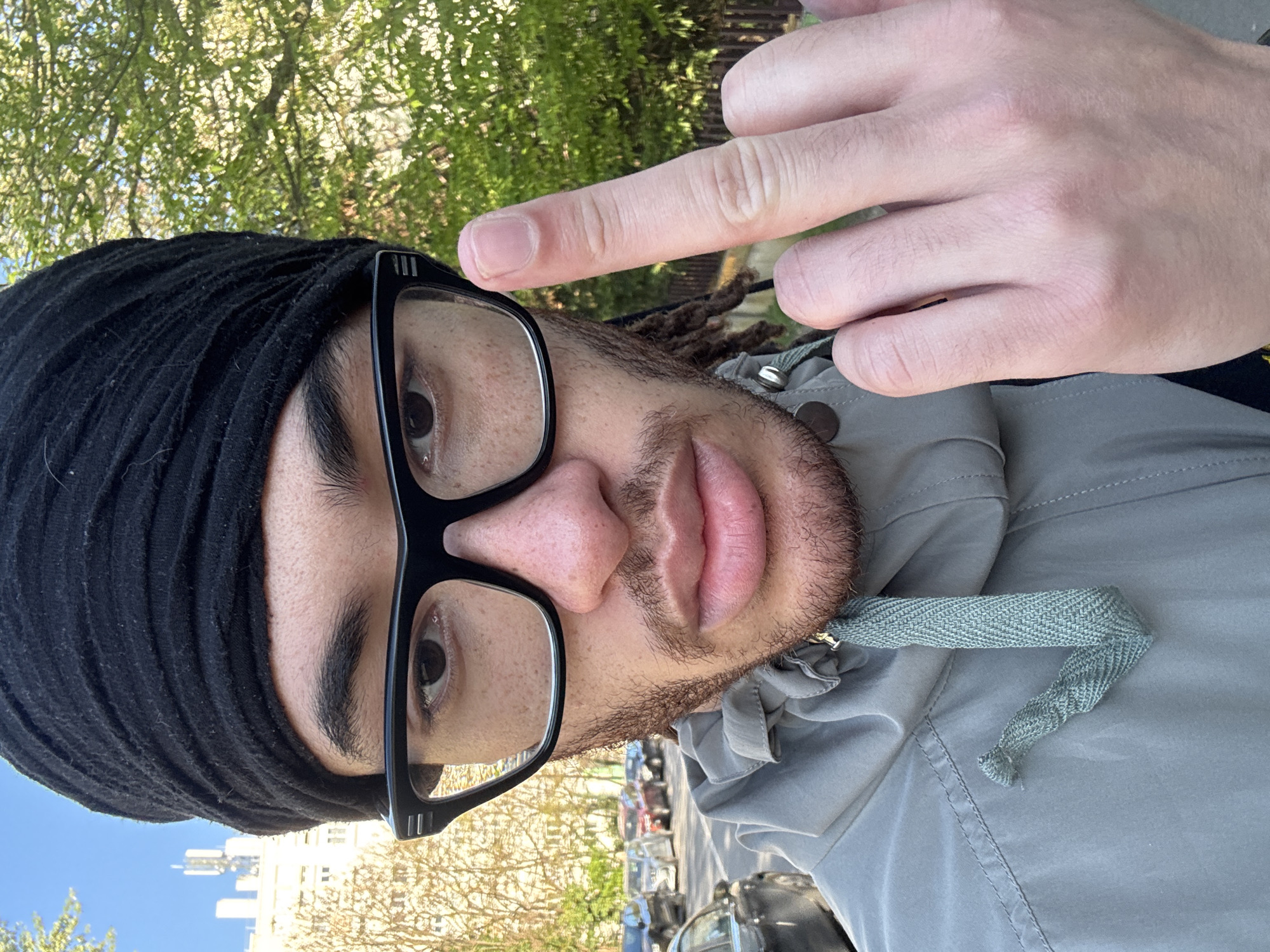A medium close-up shot depicts a man with light brown skin, wearing black-framed glasses and a black head wrap. He has a trimmed beard and some visible dreadlocks. The man is giving the middle finger to the camera. He is wearing a light grey jacket with a herringbone drawstring. The background shows trees and buildings under a bright sky. The location is identified as Les Ulis, France.