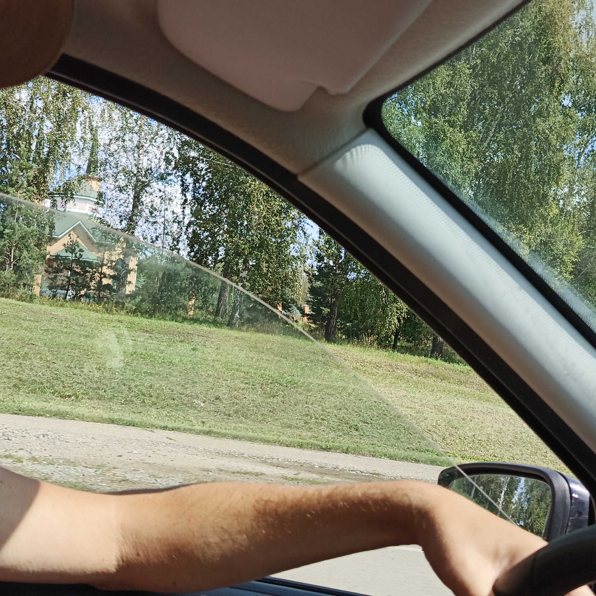 The image shows a view from inside a car, looking out the passenger side window. A person's arm and hand are visible on the right side of the frame, resting on the steering wheel or dashboard. Outside the window, there is a grassy slope with a gravel road at the bottom. In the background, there are trees and a building with a green roof and a steeple, possibly a church. The image was taken in Chishmy, Russia.