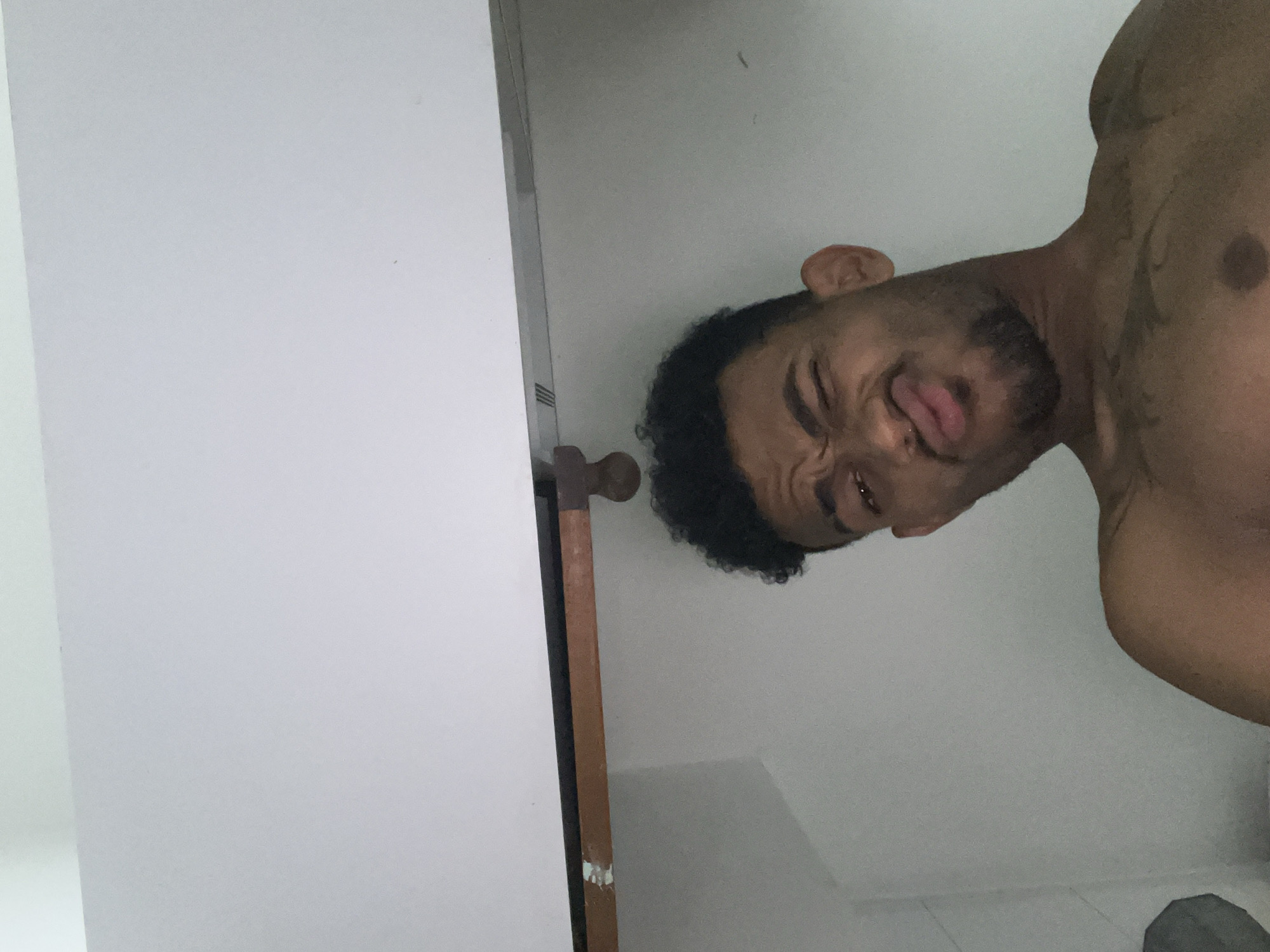 The scene takes place in the city of Porlamar, Venezuela. The image features a male individual with dark skin, short, dark, curly hair, a short beard, and a small stud nose piercing. He is captured from a low-angle perspective, looking upwards at the viewer, and displays an exaggerated facial expression, characterized by squinted eyes, a wrinkled nose, and puckered lips. The individual is bare-chested, revealing a dark-ink tattoo on his upper left pectoral muscle and a visible nipple on his right pectoral. Overhead, a white flat surface, likely a ceiling or the underside of a shelf, is visible. Immediately beneath this surface is a thin, dark horizontal line, followed by a light brown wooden beam with traces of pale greenish-blue paint on its left side. From the wooden beam, a dark, cylindrical object, possibly a cabinet handle or latch, protrudes downwards. The background behind the individual consists of a plain, light grey wall. The lighting appears even, suggesting an indoor environment.