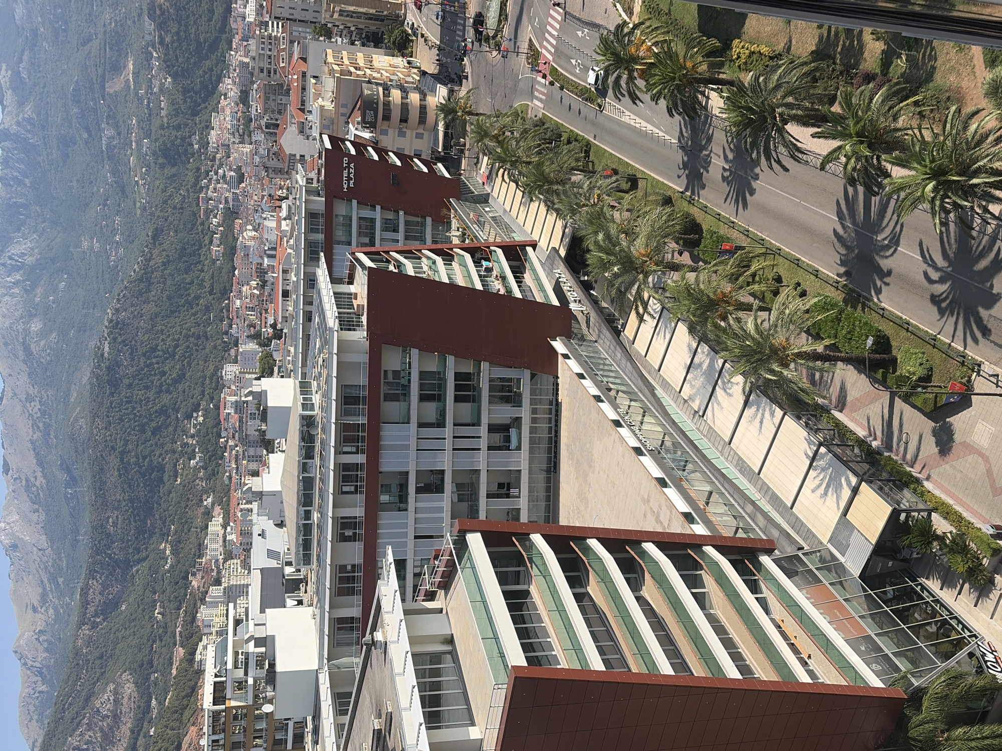 The image captures an aerial view of a modern city district, likely in Budva, Montenegro, characterized by contemporary architecture and a backdrop of lush green mountains. Dominating the foreground are several multi-story buildings with sleek, geometric designs, featuring extensive glass facades, balconies, and reddish-brown exterior panels. One prominent building displays signage for "HOTEL TO PLAZA," indicating its function as a hotel and commercial complex. Adjacent to the hotels, a "SHOPPING CENTER" is visible, with its storefronts and awnings contributing to the commercial landscape.
A paved road runs alongside the buildings, lined with palm trees that cast dappled shadows on the asphalt. The road is marked with white lines and features pedestrian crossings. Several cars are present on the road, suggesting ongoing traffic. On the sidewalks and in front of the establishments, a few individuals can be observed, their activities not distinctly discernible from this vantage point.
The urban environment transitions into a densely populated residential area with buildings of varying heights and architectural styles, many topped with red-tiled roofs, which gradually recedes towards the base of imposing, verdant mountains. These mountains form a dramatic natural boundary, covered in dense vegetation and showing signs of human habitation in the form of scattered buildings. The sky above is a clear, pale blue, typical of a sunny day. The overall scene conveys a sense of a thriving coastal city nestled between the sea (implied by the context of Budva) and mountainous terrain.