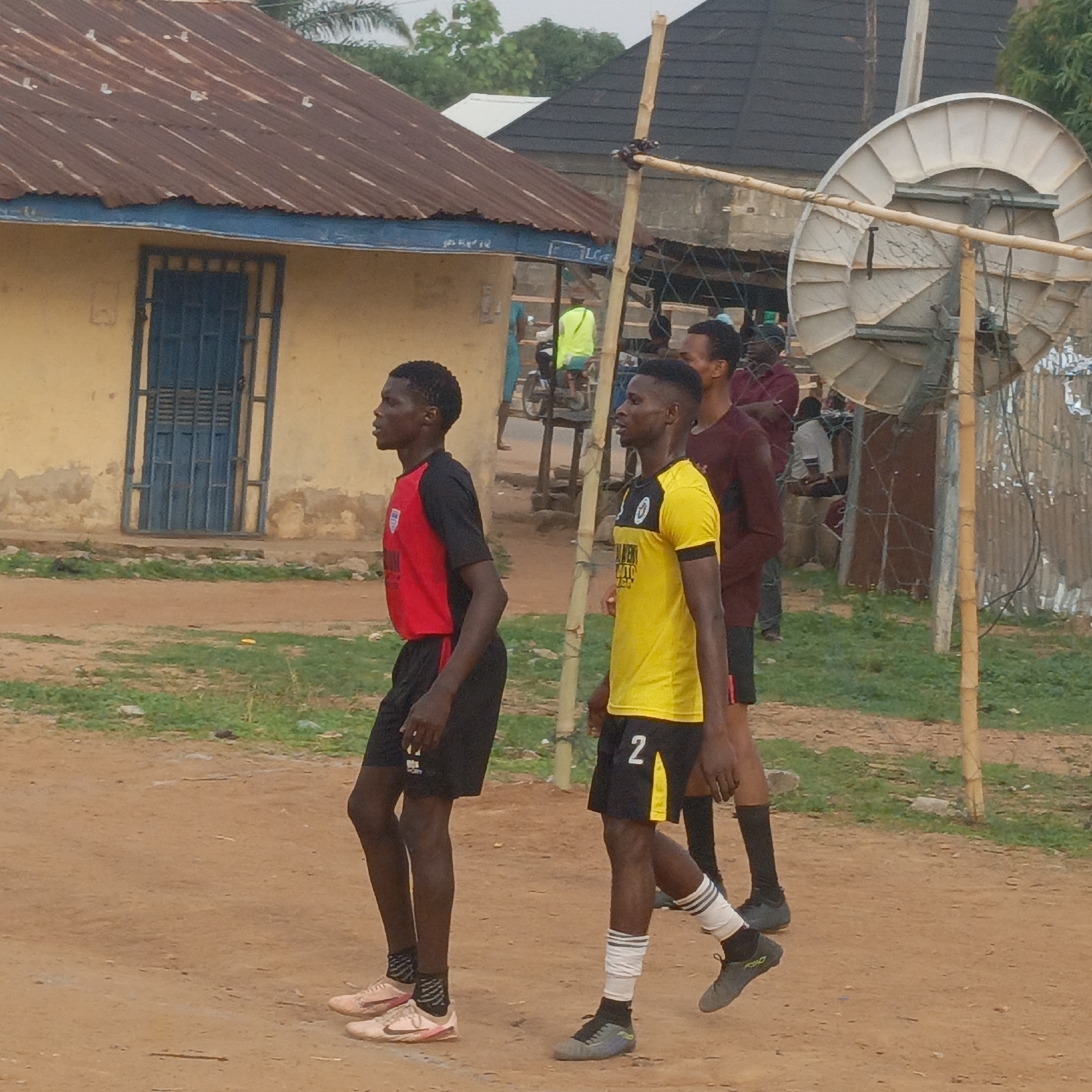 This image depicts an outdoor scene in Akwanga, Nigeria, featuring three young men, presumably footballers, in the foreground against a backdrop of local buildings and an unpaved landscape.
The ground is predominantly reddish-brown dirt, interspersed with patches of green grass. On the left, a man in a red and black short-sleeved jersey and black shorts, wearing light-colored athletic shoes with pink accents, stands in profile looking right. Central to the frame, another man in a yellow and black short-sleeved jersey and black shorts with the number "2" visible, also faces right. He wears black and white striped socks and dark athletic shoes. Partially visible on the far right, a third man wears a maroon long-sleeved shirt and dark shorts. Their attire suggests they are either practicing, playing, or resting from a football match.
In the midground, an improvised structure made of bamboo poles, possibly a goal post, is visible, and a large, grey satellite dish is mounted nearby. The background features several buildings, including one with a yellow-painted wall, a rusty corrugated iron roof, and a blue metal door/gate. Other less distinct structures and individuals, including one on a motorcycle, are visible further back.
The scene occurs during daytime under what appears to be an overcast or hazy sky, as indicated by the soft, diffused light and lack of strong shadows. The general atmosphere is calm, and the individuals appear relaxed. The visual cues, including the building styles and ground cover, align with the provided context of Akwanga, Nigeria.