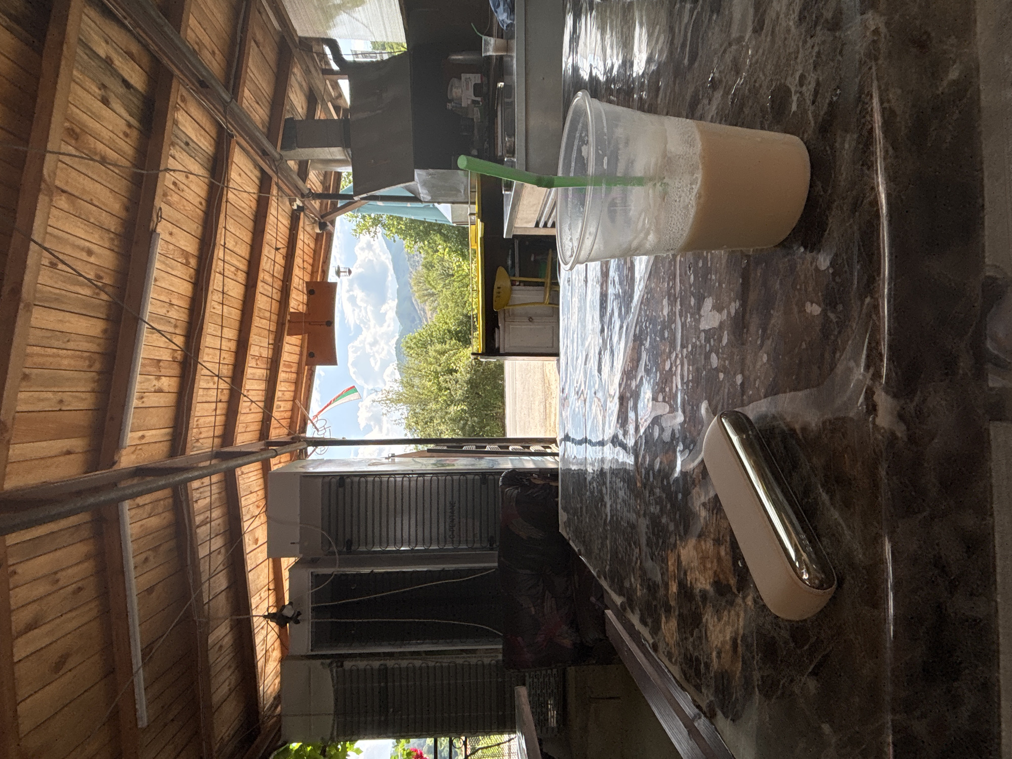 The image captures an outdoor seating area, likely a cafe or restaurant, with a wooden-slatted roof supported by metal beams. In the foreground, a marble-patterned table is covered with a clear plastic sheet, showing some spills. A disposable plastic cup containing a light brown beverage with a green straw sits prominently on the table. Next to it, a metallic gold and beige object, possibly an electronic device, rests on the table.

In the mid-ground, industrial-looking refrigerators are lined up against a wall. Further back, a striped sofa is visible. Beyond the seating area, an open-air structure houses kitchen equipment, including a grill. A yellow chair is placed near a counter. The background reveals a rural landscape with lush green trees and hills under a cloudy sky. A flag, appearing to be the flag of Bulgaria, flutters in the distance. The scene suggests a casual, perhaps slightly rustic, dining or resting spot. It is located in the city of Mesta, Bulgaria.