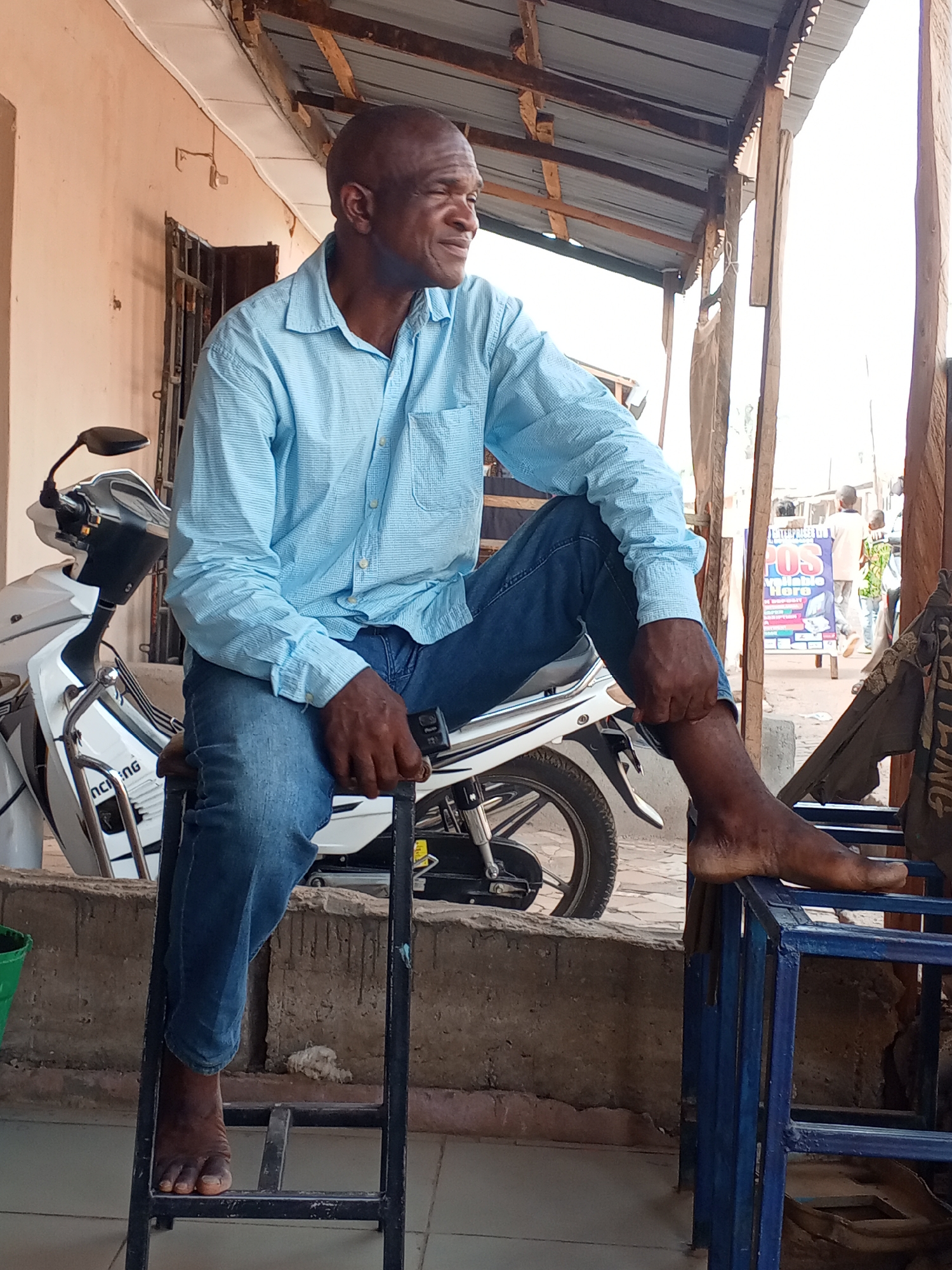 In Sabon Tasha, Nigeria, a bare-footed Black man, wearing a light blue long-sleeved button-up shirt and jeans, sits outside on a metal frame stool. One of his feet rests on a blue metal structure. Behind him is a white scooter parked next to a building with a beige exterior wall. In the background, a sign is visible, and other people walk by. A corrugated metal overhang is above the seated man.