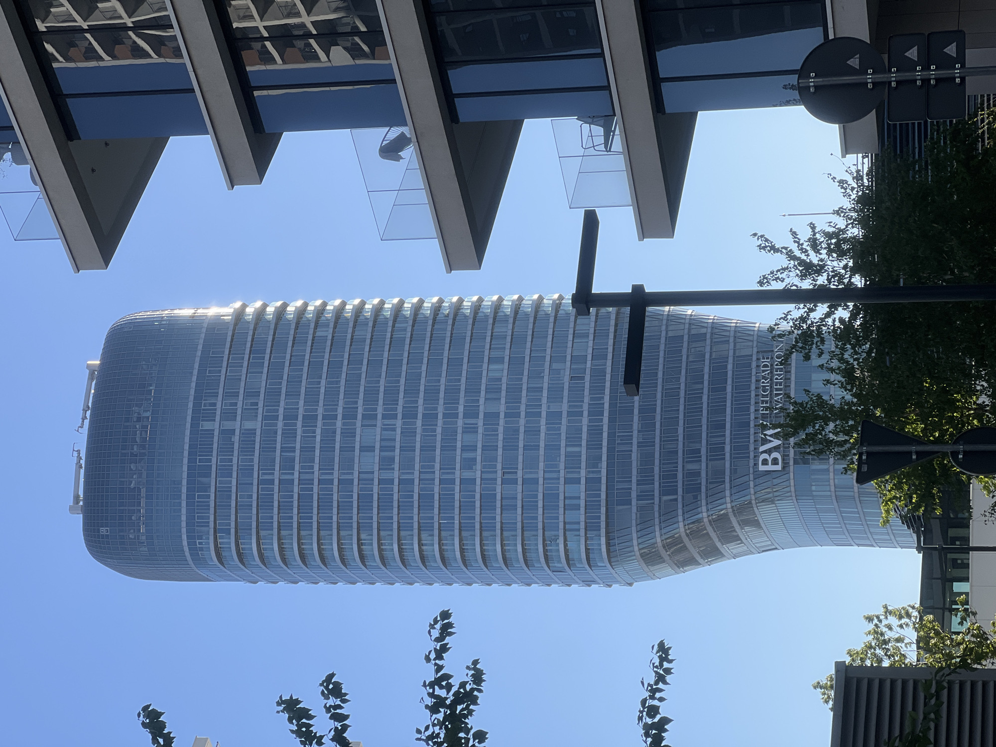 The image is an upward, slightly distorted view of the BW Belgrade Waterfront building in Belgrade, Serbia. The building is a curved, cylindrical high-rise with numerous rows of windows, and the "BW BELGRADE WATERFRONT" logo is visible on its side. Below the building, there is sparse foliage. Above, the image is framed by the architectural details of another building, including cantilevers and glass panels. A street sign is also visible, partially obscured by foliage. The sky is clear and blue.