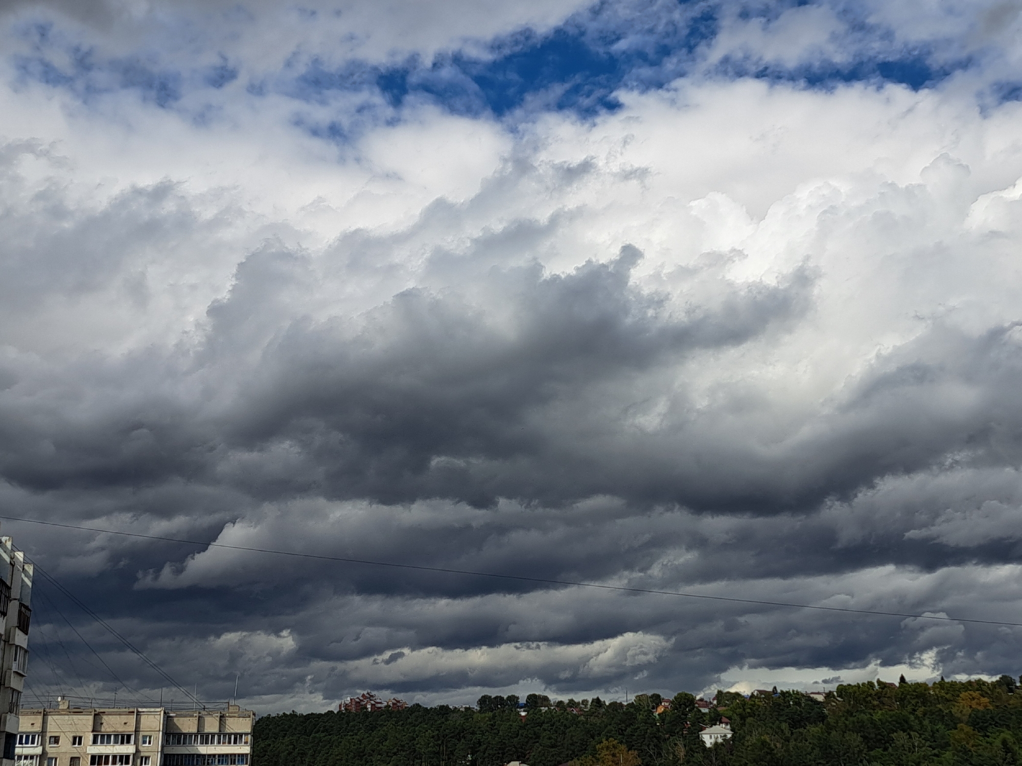 The image depicts a cloudy sky over a city. In the foreground, two apartment buildings are visible on the left and center. The building on the left is partially visible, showing windows and balconies. The building in the center is more fully in view, also with numerous windows. A thin, dark line, likely a power line, stretches horizontally across the middle of the frame. Below the power line, a dense line of trees forms a dark green band across the landscape, with some rooftops of houses visible peeking through the foliage. The sky is filled with a variety of clouds, ranging from bright white, puffy formations at the top to dark, heavy, and ominous storm clouds lower down. Patches of blue sky are visible in the upper portion of the image. The overall impression is of a cloudy, possibly overcast day, with the looming presence of storm clouds suggesting an impending change in weather. The architecture of the buildings and the type of trees are consistent with an urban environment in Russia, and specifically in Irkutsk.