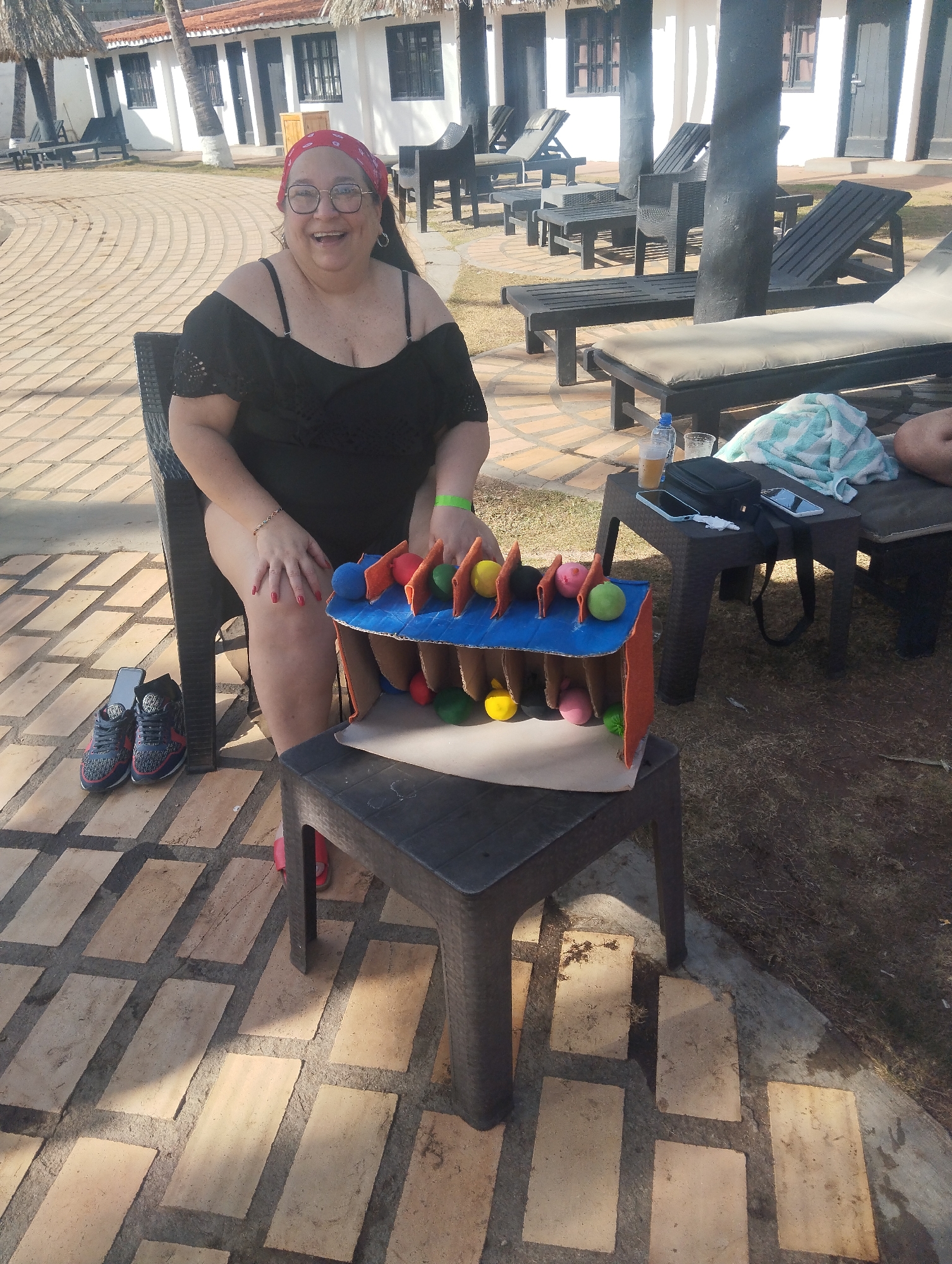 The image shows a woman seated in a black chair outdoors in Porlamar, Venezuela. She is wearing a black swimsuit and a red bandana. She has glasses on and is smiling. Next to her, there is a small black table with a homemade game made of cardboard and colorful balls. The game appears to be a vertical maze-like structure. Behind her are several lounge chairs and tables on a tiled patio. In the background are white buildings with dark brown roofs and doors. Other accessories on the chair include sneakers on the floor beside her. On the adjacent table are a cellphone, towel, beverage and purse.
