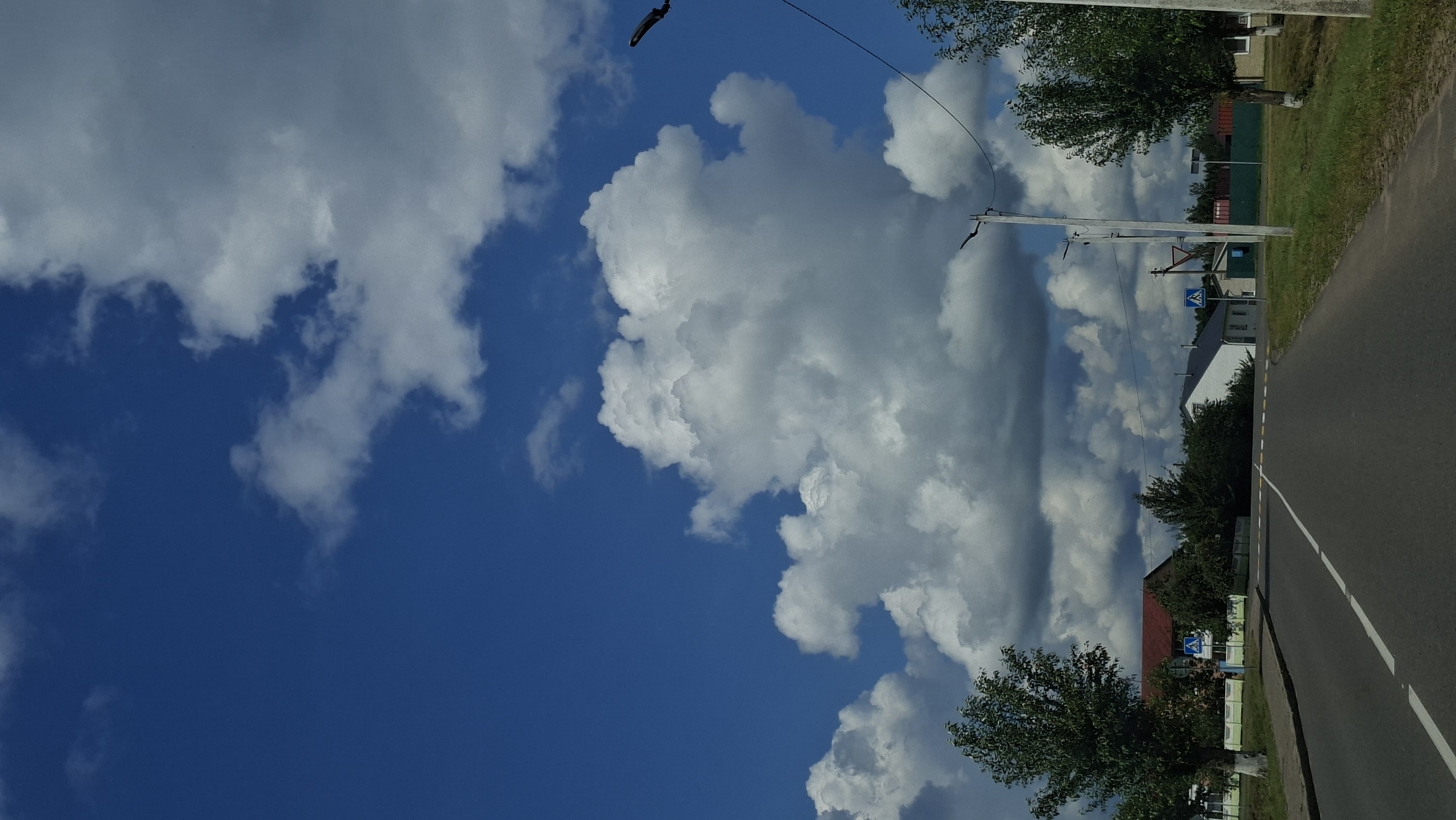 The image captures a bright, sunny day with a vast, vivid blue sky filled with large, fluffy white clouds. A paved road stretches into the distance, with white lines marking its lanes and a yellow speed bump crossing it. On either side of the road, green grass and trees are visible. Several houses, some with red roofs and others white, are situated along the road. A pedestrian crossing sign is present. In the foreground, a single tree stands to the left, while to the right, another tree grows near a utility pole. The scene appears to be a residential area in the city of Myadzyel, Belarus.