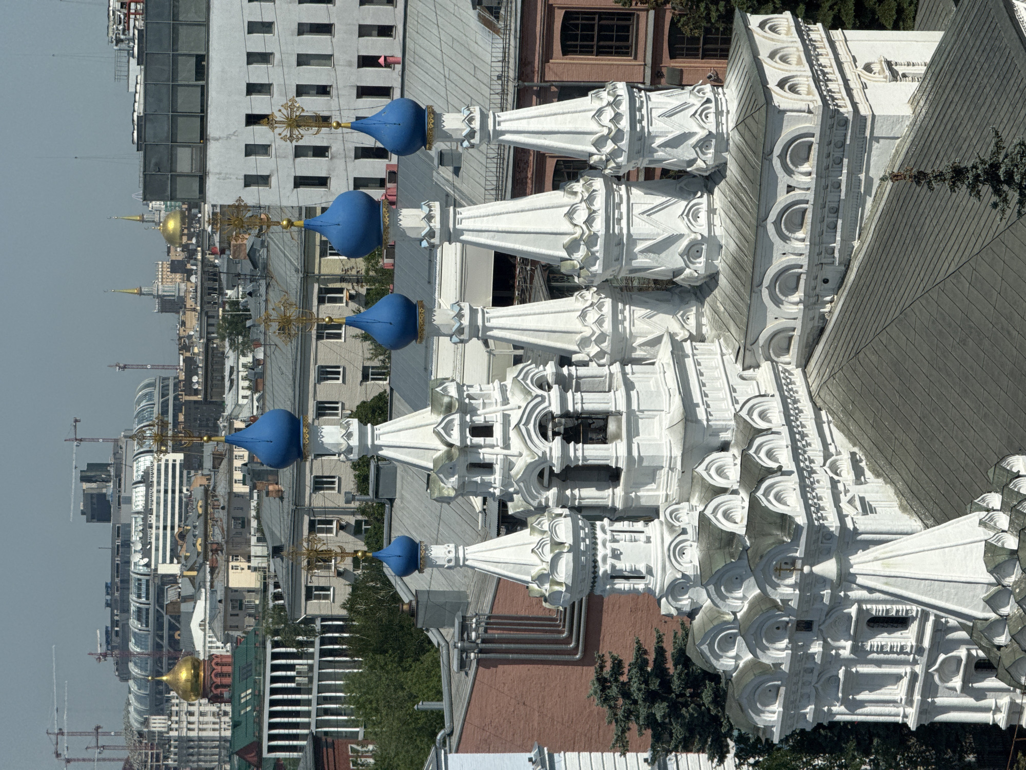 The image shows an elevated view of the Church of St. Nicholas in Khamovniki in Moscow, Russia. The church is prominently featured with its white facade, intricate architectural details, and blue domes. The cityscape behind the church includes various buildings, from older, shorter structures to taller, modern high-rises, and construction cranes suggesting ongoing development in the area. The overall atmosphere is urban and densely populated.