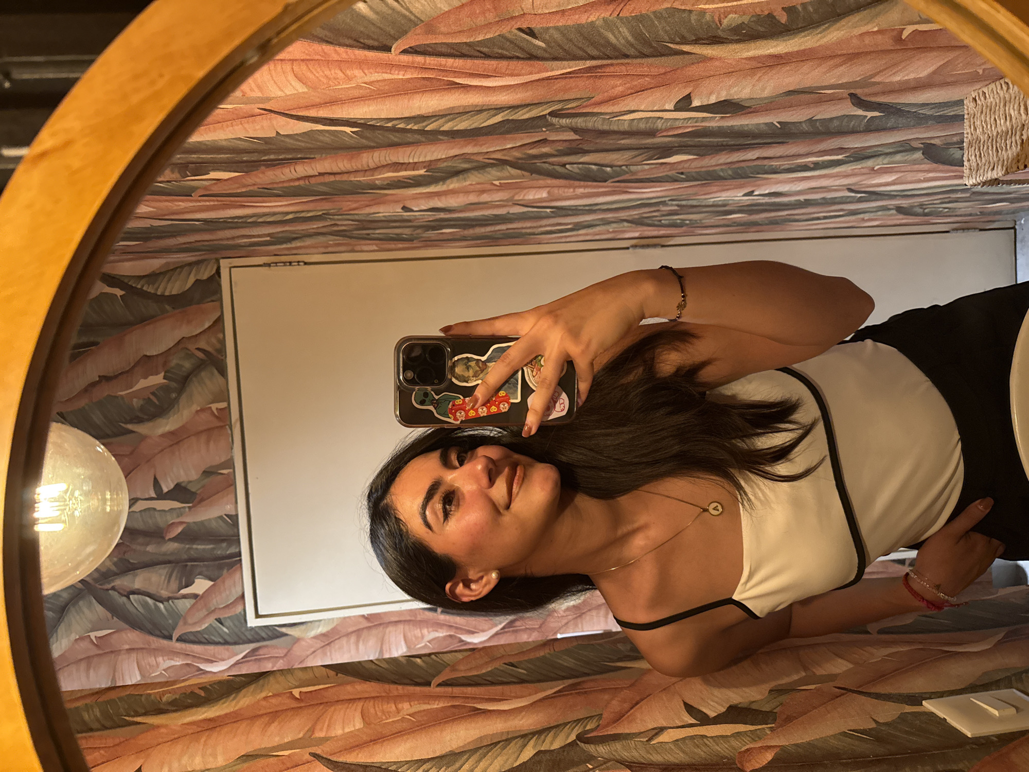 A woman is taking a selfie in a mirror. The mirror has a wooden frame and is reflecting a bathroom scene. The background wallpaper features a repeating pattern of stylized leaves in shades of brown, tan, and green. A light fixture is visible on the left, partially obscured by the mirror's frame. The woman is wearing a white top with black trim and a necklace. She's holding a smartphone in her left hand, and its case is adorned with stickers. The room includes a white framed rectangle, possibly a picture or a cabinet door. The location is not explicitly stated in the image, and the city of Puebla, Mexico, cannot be identified from the image alone.