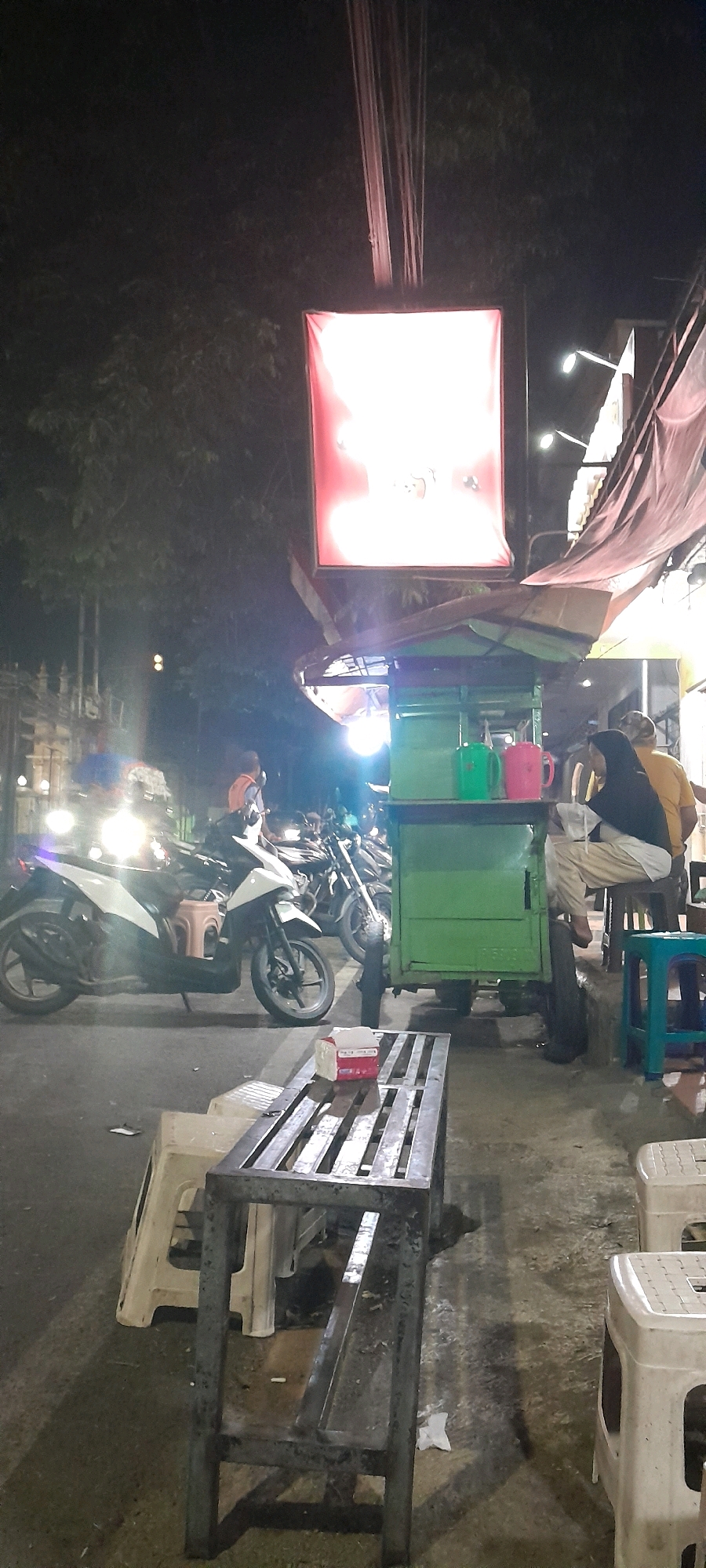The image depicts a nighttime scene at an outdoor food stall in Rogojampi, Indonesia. A green food cart with a metal roof and awning is positioned in the center of the frame, displaying two mugs, one green and one pink, on its counter. Behind the cart, a person wearing a yellow shirt and a black headscarf is seated on a stool, while another person in a yellow vest is standing.
In the foreground, a metal bench with a slatted surface sits on the pavement. To the left of the bench, a white plastic stool is visible. Several motorcycles are parked on the street in the background, with their headlights illuminated. A large, illuminated red sign hangs above the food cart, casting a soft glow on the surroundings. The pavement is wet, suggesting recent rain or a damp environment. The overall atmosphere is that of a bustling street scene at night, typical of a local food stall in Indonesia.