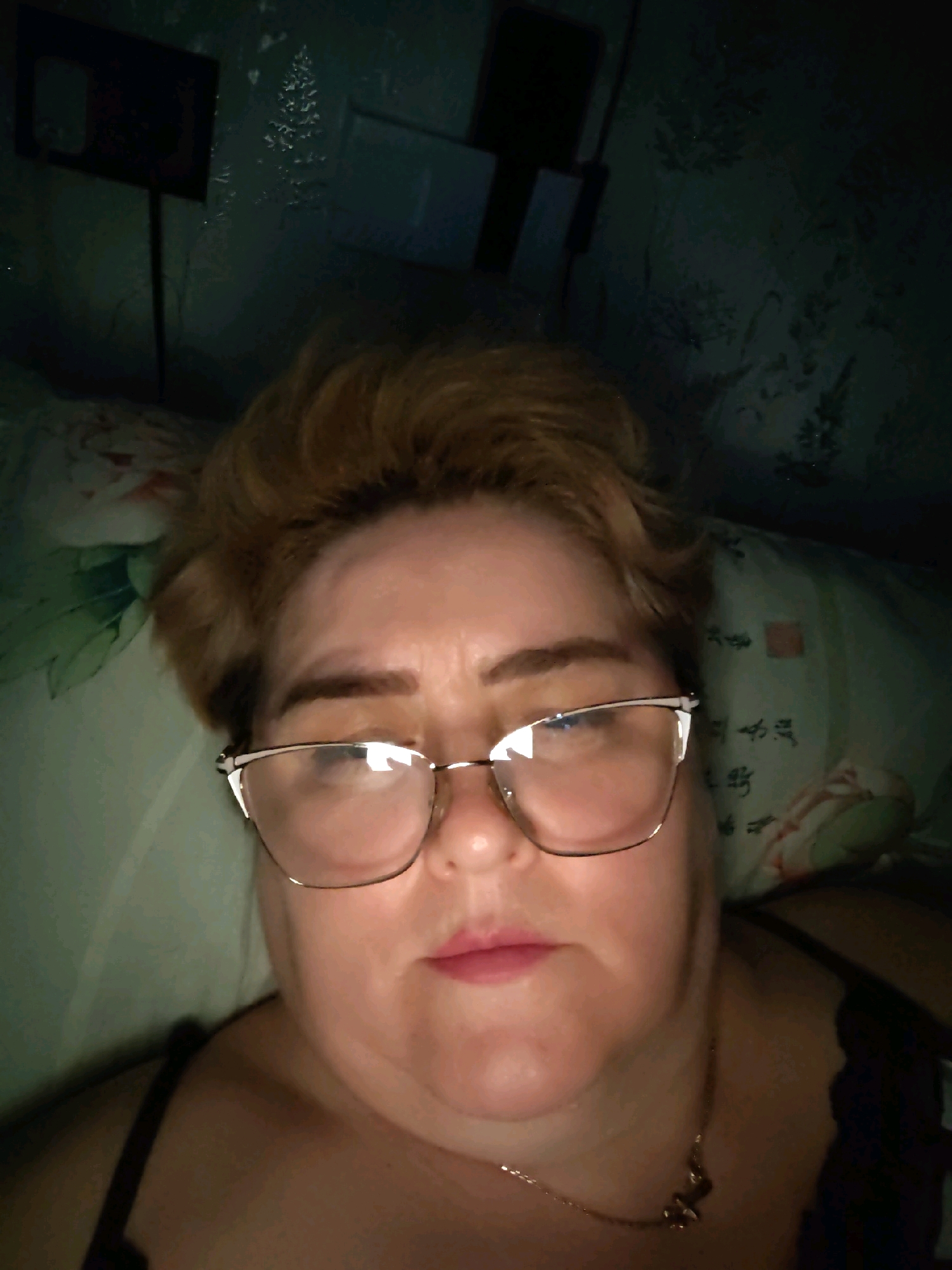The image is a selfie taken indoors, likely in a bedroom, as indicated by the pillows and bedding. The subject is a woman wearing glasses and a necklace. The lighting is dim, and the background appears to be a patterned wallpaper. There are no identifiable landmarks or context within the image to suggest a specific location within the city of Shchyolkovo, Russia, or any other city. The scene depicts a close-up of the woman's face and upper body.