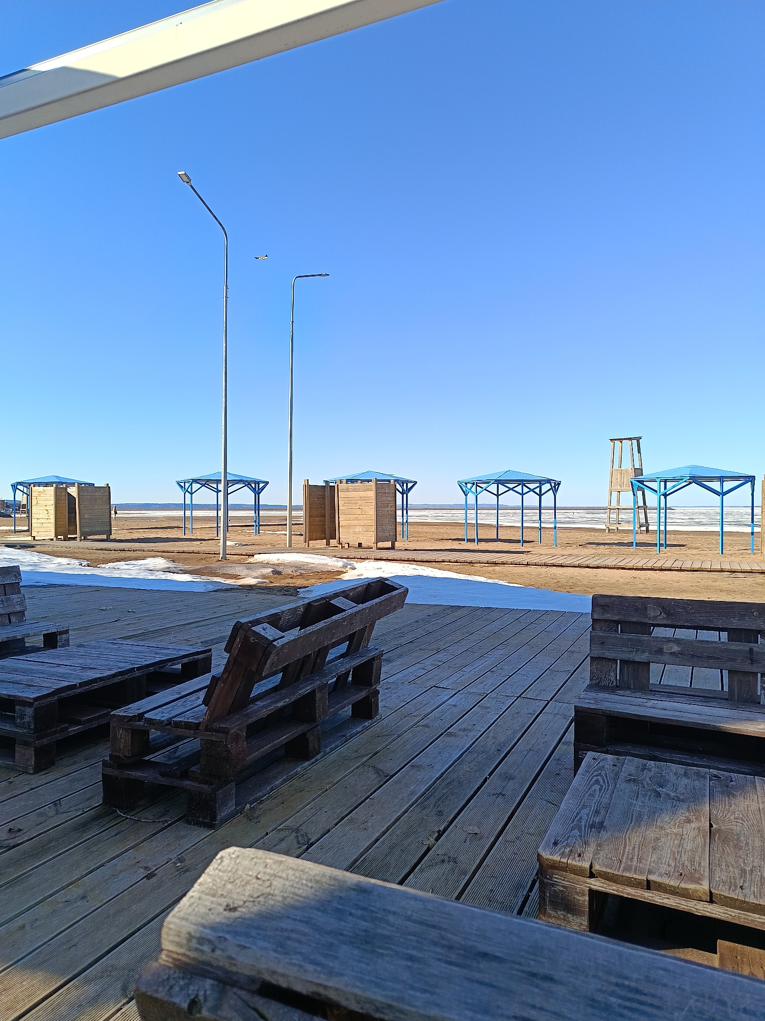 The image depicts an outdoor seating area, constructed from wooden pallets, located on a boardwalk. In the background, the scene extends to a beach under a clear blue sky. On the beach are several blue-roofed structures and wooden changing stalls. Two streetlights are visible against the sky. Patches of snow are interspersed on the boardwalk and beach. The photograph was taken in the city of Chaikovsky, Russia.