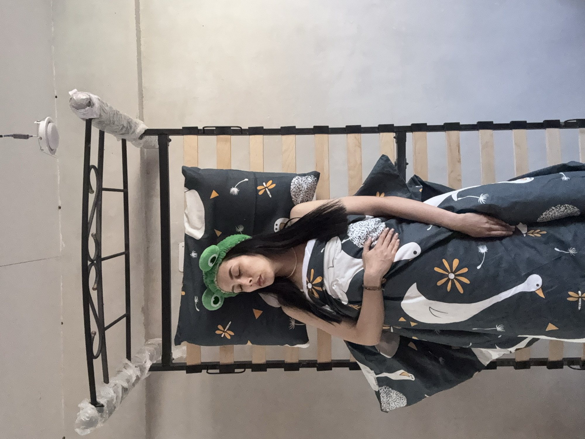 A woman is lying asleep in a bed. She is wearing a green frog-shaped headband. The bed has a black metal frame and wooden slats. The bedding is patterned with white birds and yellow flowers. The location is not identifiable as Votkinsk, Russia based on visual information.