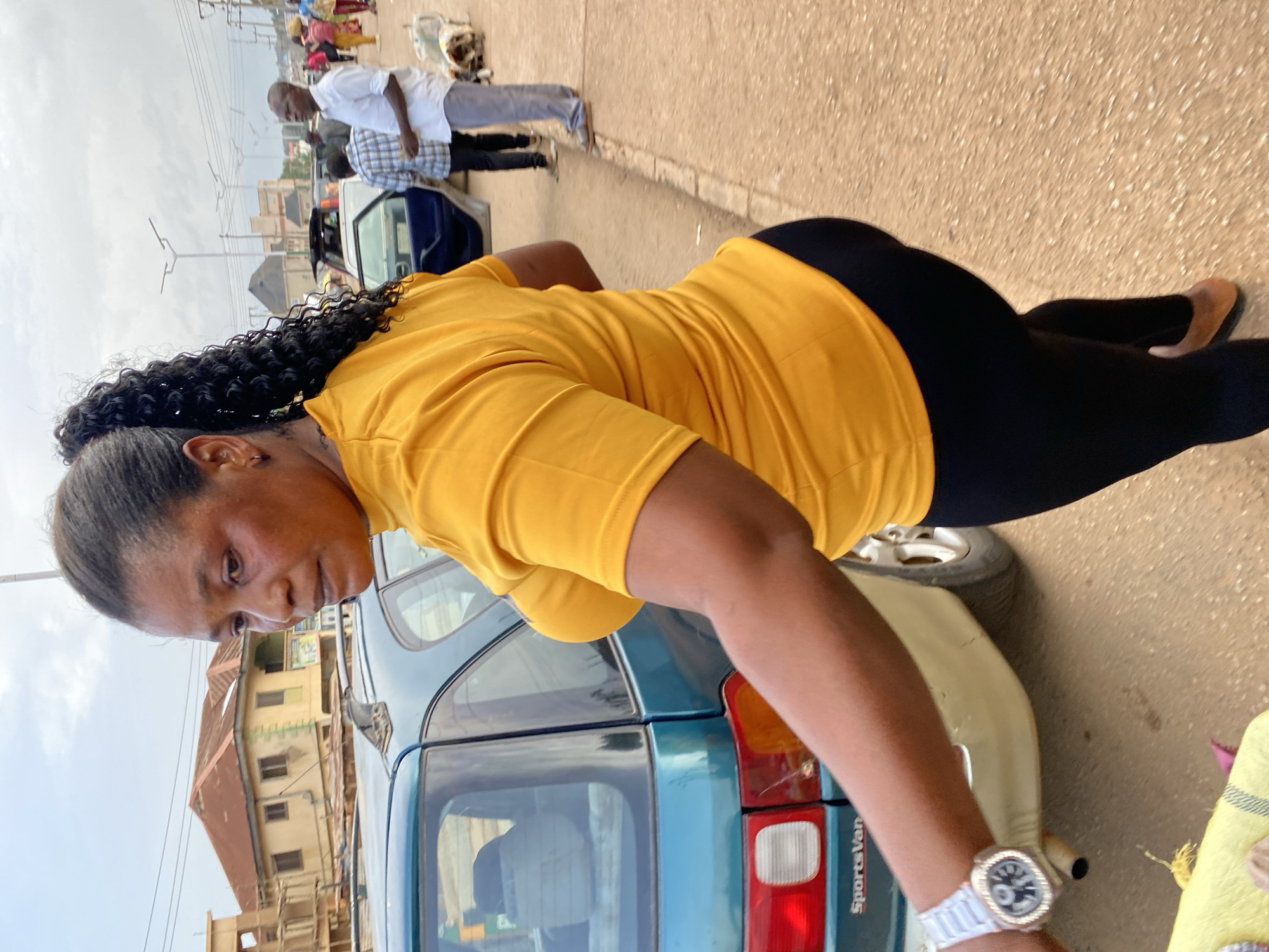 In Bukuru, Nigeria, a woman with dark skin and a ponytail of curly dark hair is seen from the side. She is wearing a yellow t-shirt and black leggings. She has a watch on her left wrist. The woman is standing next to a teal colored car. In the background, there are buildings, streetlights, and other people. The ground is a dusty, reddish-brown color. The sky is overcast and gray.