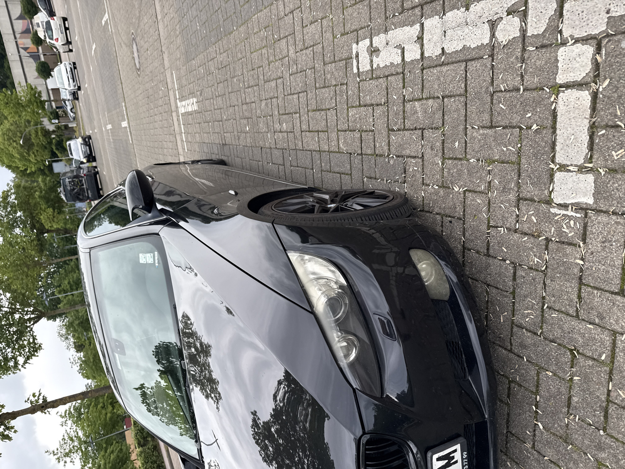 This image captures a black BMW 3-series parked on a paved surface in what is identified as Delmenhorst, Germany, during daylight hours under an overcast sky.
The primary subject is the front three-quarter view of a black BMW, likely an E90-series, positioned towards the right of the frame. Its glossy black paint reflects the overhead sky and surrounding trees. The vehicle features multi-spoke black alloy wheels, a distinct BMW kidney grille, and dual circular projector headlights, which show some minor haziness. A German license plate, partially visible at the lower left of the front bumper, displays "M" followed by the numbers "3 77 99". The windshield has a rain sensor/camera module visible at the top center and several stickers on the passenger side.
The ground is paved with rectangular grey interlocking concrete pavers laid in a patterned arrangement. White painted lines, indicating parking spaces, are visible, showing signs of wear and partial fading. Scattered across the paved surface are numerous light-colored, elongated leaf-like debris.
In the mid-ground and background, additional vehicles are present on a similar paved surface. These include a black commercial van with a bicycle rack attached to its rear, a white hatchback (possibly a Volkswagen Up!), a white sedan, and several other vehicles in the distance. To the far left and in the upper background, multiple deciduous trees with green foliage are visible, indicating a vegetated area. A concrete wall or structure with a faint geometric pattern is also visible in the background, behind the parked cars. No individuals are present in the frame. The overall light suggests an overcast or cloudy day.