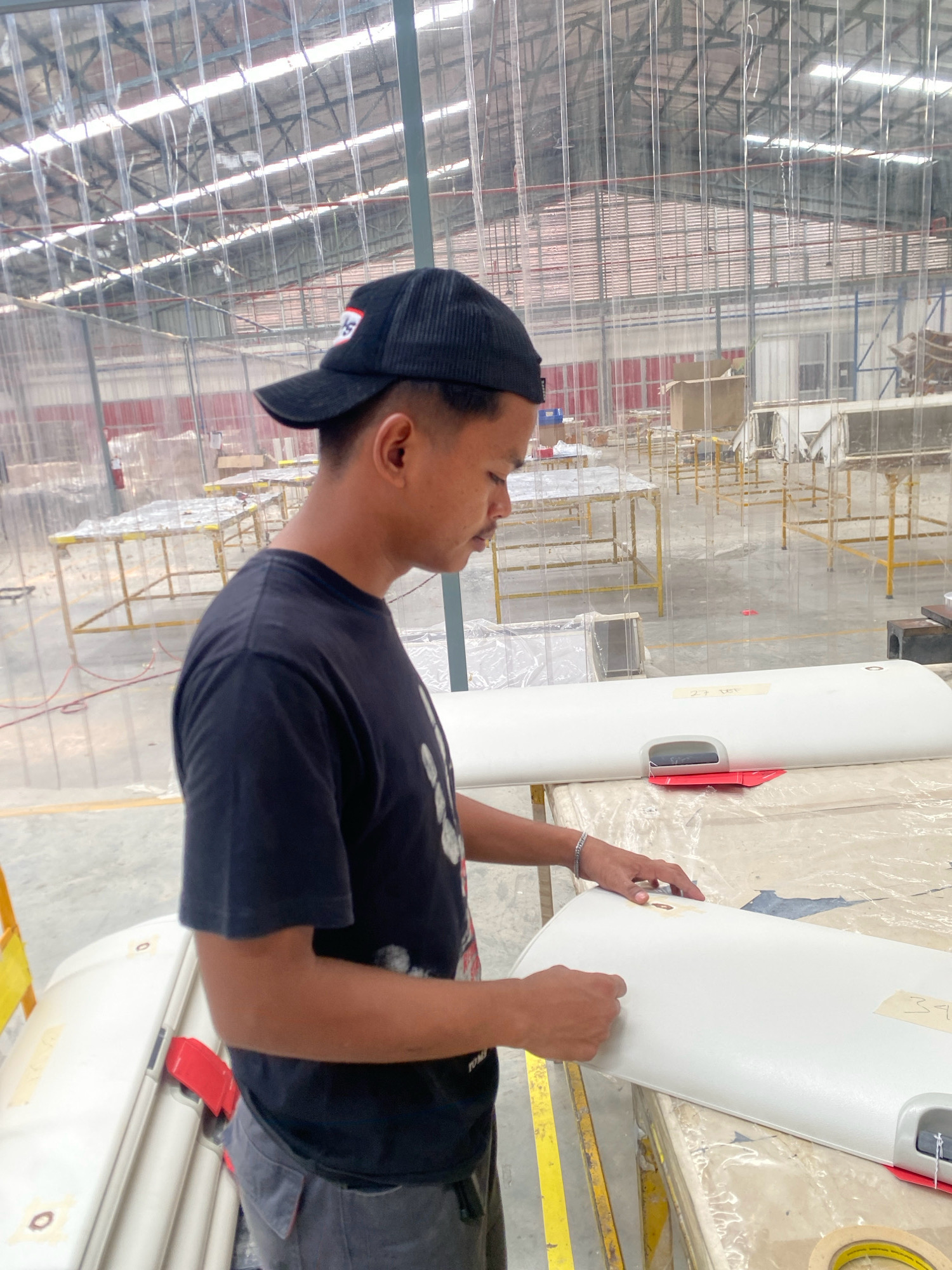 An outdoor, medium shot captures a man wearing a black t-shirt, grey pants, and a black baseball cap working on a large, white, curved object in what appears to be a factory or workshop environment in Batam, Indonesia. The man is standing and looking down at the object, with his hands actively engaged in some form of assembly or inspection. The object itself is smooth and white, with a handle-like protrusion and some visible tape or labeling. The background reveals a large, open space with what looks like a corrugated metal roof and translucent, vertical partitions, suggesting a manufacturing facility. Yellow metal shelving units and other white objects are visible in the distance, contributing to the industrial setting. The lighting appears to be natural, coming from above, and the overall atmosphere is one of focused work within a production area.