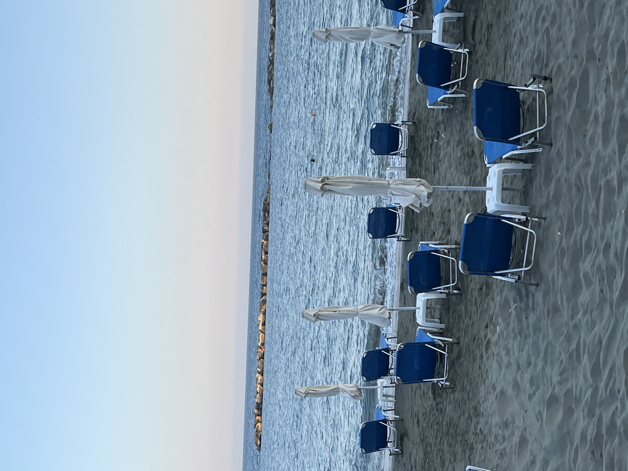 The image depicts a serene beach scene in Agios Tychona Community, Cyprus. In the foreground, several blue lounge chairs are arranged on the dark sand, some with white umbrellas covering them. The chairs are empty, suggesting a quiet moment before or after a busy period. The ocean stretches out behind the beach, its surface rippled by gentle waves. A man-made breakwater, constructed from large stones, runs parallel to the shoreline in the distance, breaking the monotony of the sea. The sky above is a soft gradient of pale blue, transitioning to a lighter hue near the horizon, indicating either early morning or late evening. The overall atmosphere is tranquil and peaceful, characteristic of a Mediterranean coastal resort.