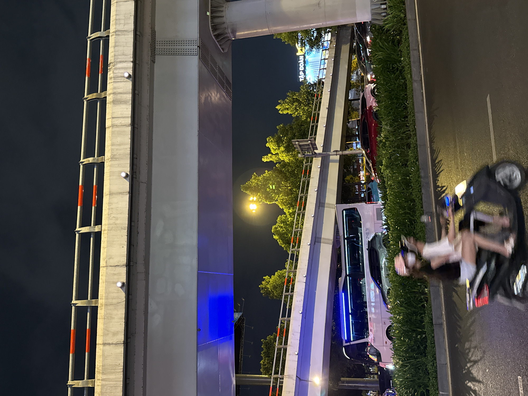Observed at night in Thủ Đức, Vietnam, a concrete overpass dominates the scene. A ladder extends up the side of the overpass, marked with orange and white stripes. A sign illuminated with the Vietnamese text "TẬP ĐOÀN" is visible beyond the overpass, surrounded by trees. Below the overpass, traffic is moving, including a large bus with blue LED accents. A motorcycle with two passengers is blurred in motion. The environment includes lush greenery adjacent to the road.