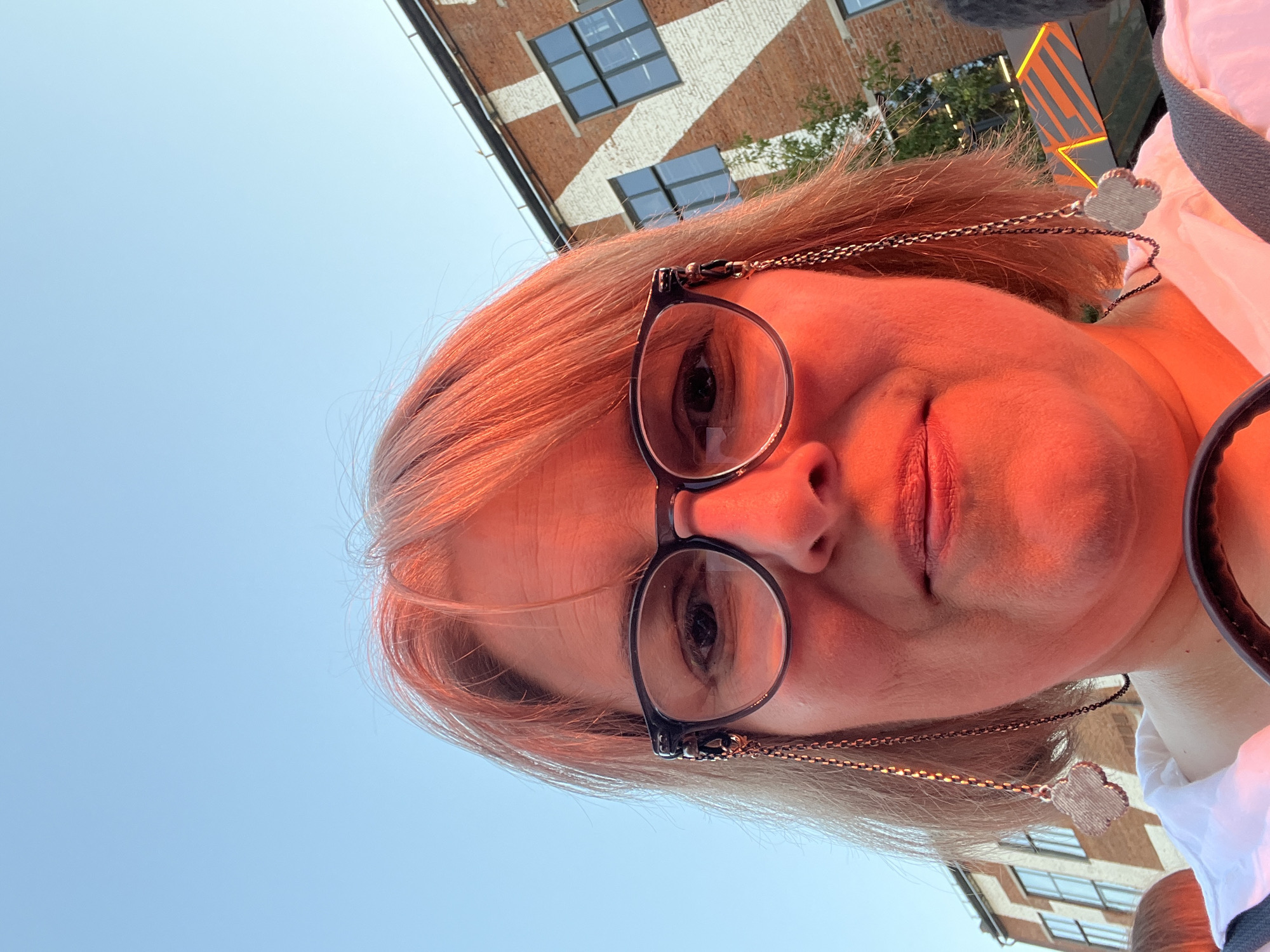 A woman with short, reddish-blonde hair, wearing glasses with decorative chains, is pictured from a low angle. She is illuminated by a strong red light, likely from a sunset or artificial source. Behind her, a brick building with distinctive white zigzag patterns and windows is visible, set against a clear blue sky. The context of the location in Khimki, Russia is provided.
