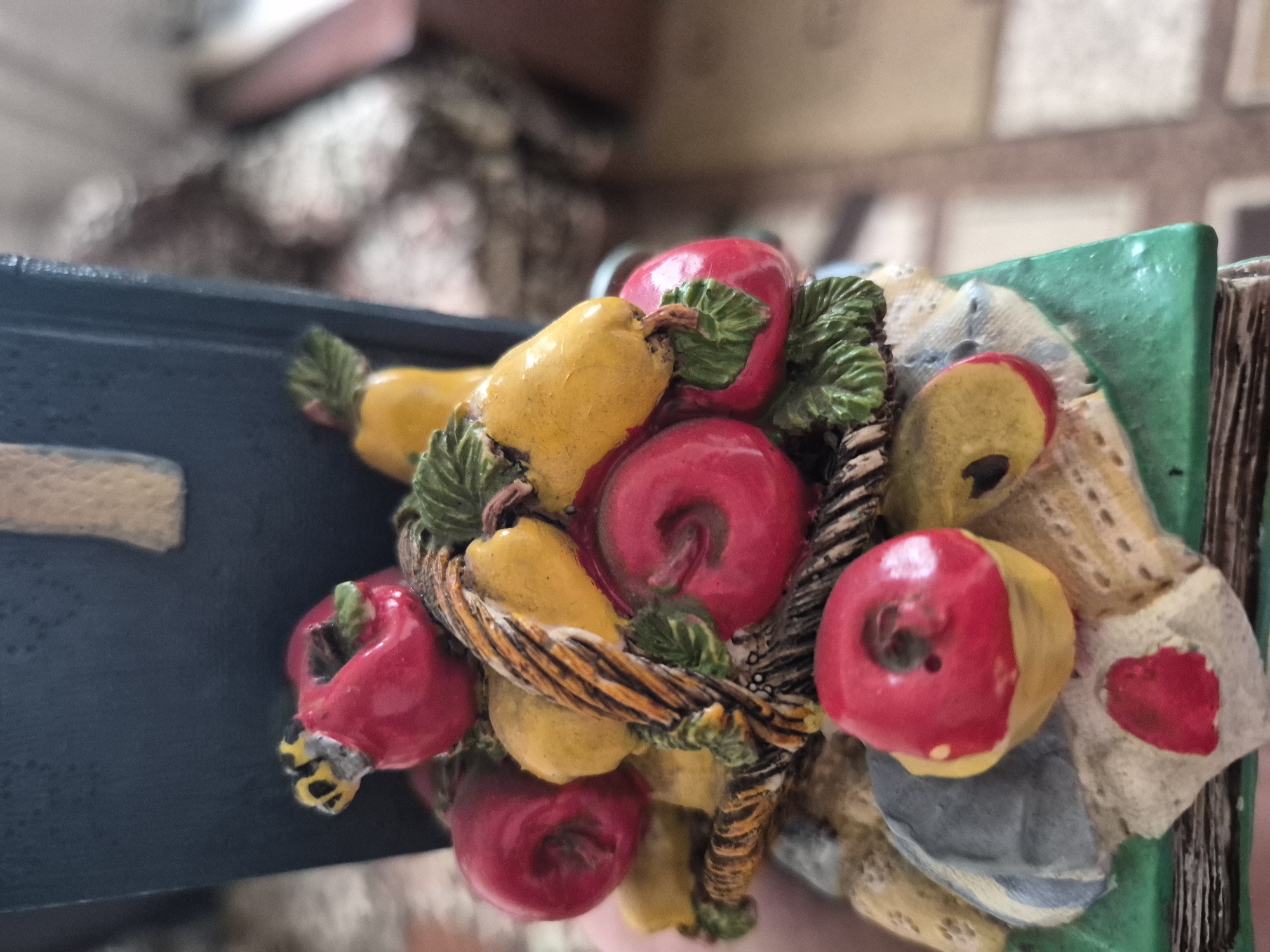 The image shows a close-up of a decorative item featuring a basket overflowing with artificial fruits, primarily red apples and yellow pears, nestled amongst green leaves. A small bumblebee figurine is perched on one of the apples. The basket itself is made of a brown, woven material. The fruits and basket are placed on a base that resembles an open book with pages and some folded fabric. The background is blurred, showing a room with patterned upholstery and wooden furniture. This item is not located in Vanadzor, Armenia; it is a decorative collectible.