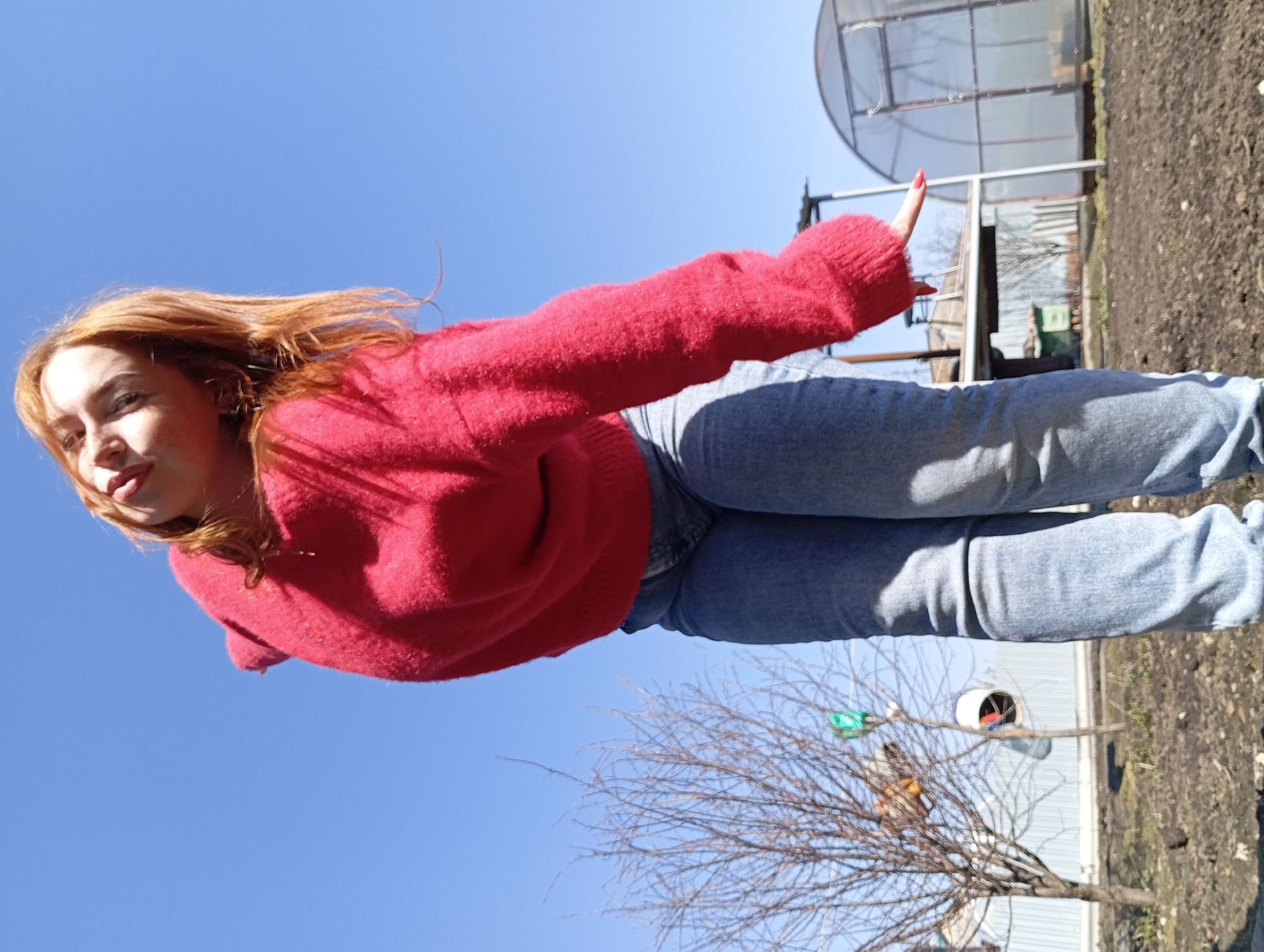A red-haired woman wearing a red sweater and blue jeans is outdoors. She has her left arm extended with her index finger pointing upward. The background includes a blue sky, a bare tree, a greenhouse-like structure, and a building. The location is Yurgamysh, Russia.