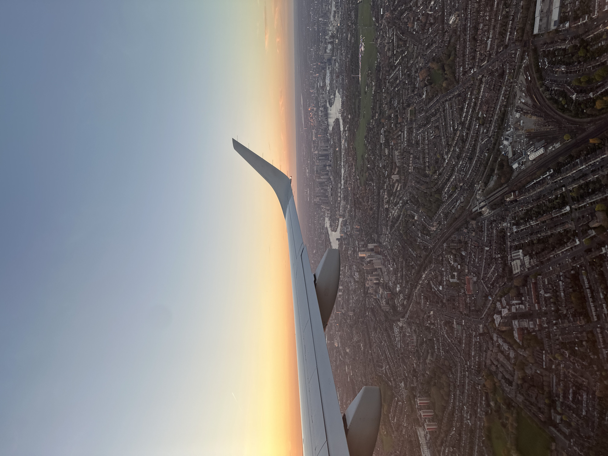 The image depicts an aerial view of London, United Kingdom, taken from an airplane. The left side of the image shows the wing of the plane against a gradient sky transitioning from blue to orange, suggestive of either sunrise or sunset.
The right side of the image displays a densely populated urban landscape. This area contains a network of residential streets lined with buildings, interspersed with green spaces. One prominent feature is the Serpentine Lake in Hyde Park, a distinctive landmark in central London. Other visible parks include Kensington Gardens and Regent's Park. The architectural style and layout suggest a primarily residential area, with occasional commercial or institutional structures. The image does not display people or actions.