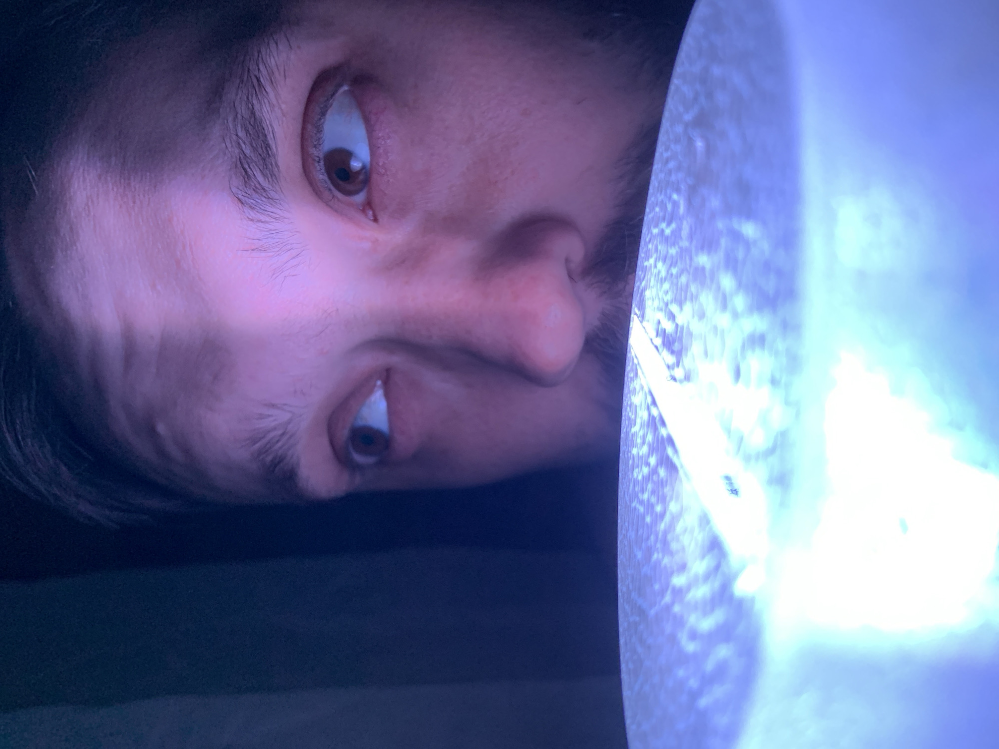 A close-up, low-angle, indoor shot shows a Caucasian male with brown hair and eyes, and a beard, looking to his left. A bright, white light illuminates the bottom left portion of the frame, creating a reflective surface that blurs the lower half of the man's face. The lighting casts a pinkish hue on the man's skin and forehead, with a distinct shadow line across his forehead. The background is dark and out of focus. There is no discernible information in this image that would allow for a definitive location in Chaikovsky, Russia.