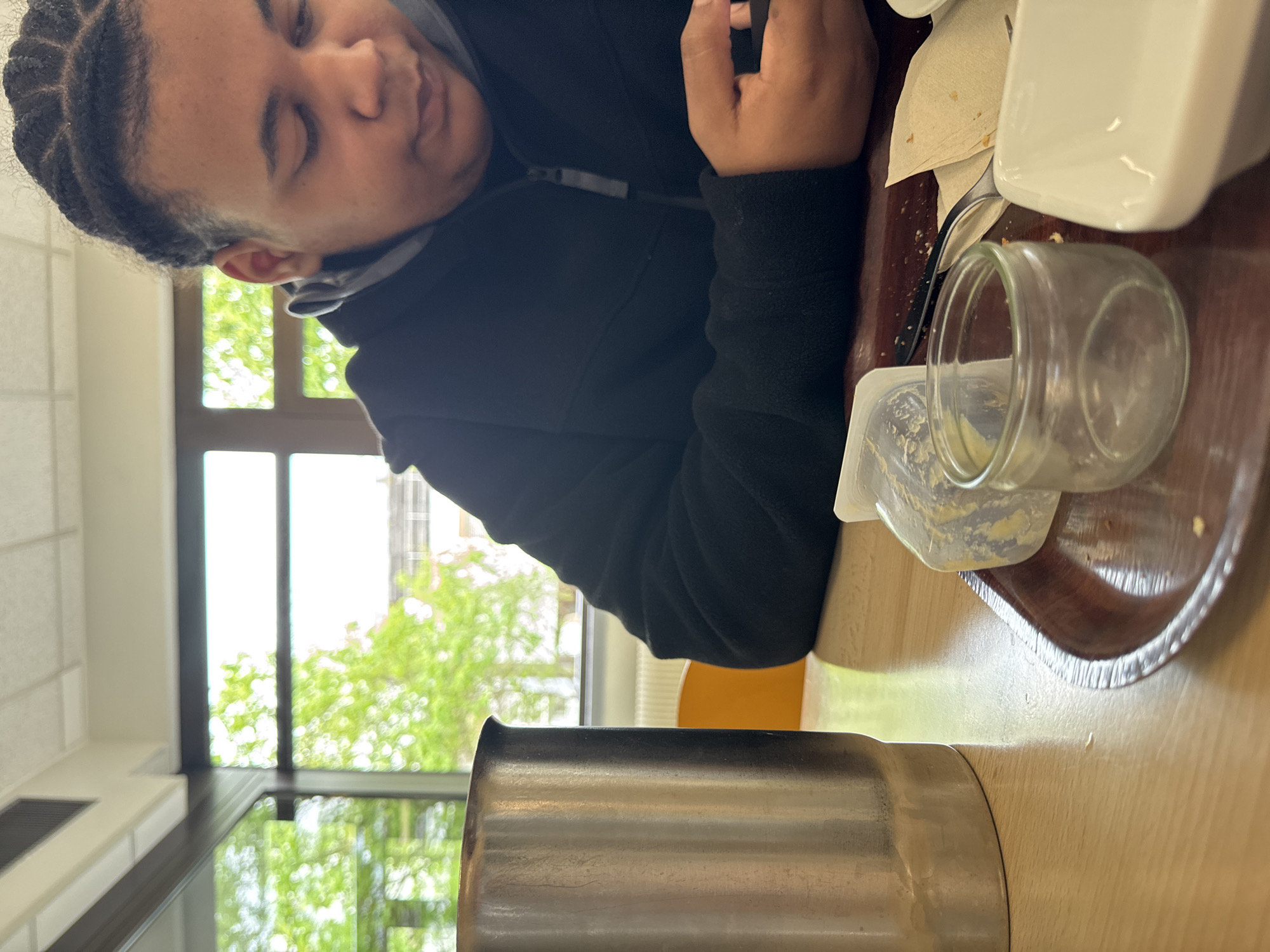 The image shows a person seated at a table in an indoor environment, potentially a cafeteria or dining area. The individual has dark hair in braids and is wearing a black jacket. Their eyes are closed, possibly resting. On the table, there's a metal container, a small glass jar, a white square dish, a napkin, and a tray. Outside the window, greenery suggests a park or garden-like setting. The location is Schiltigheim, France.