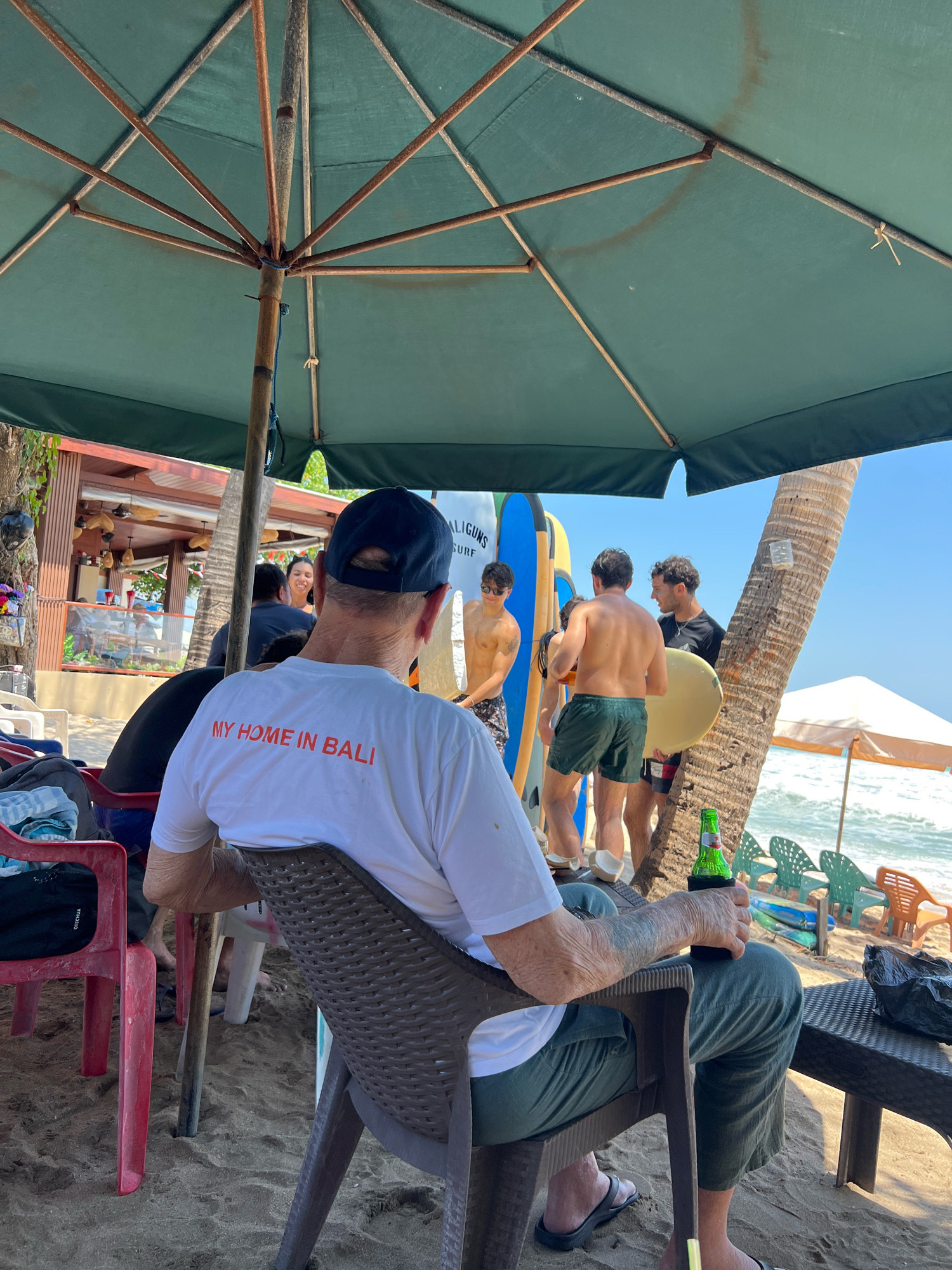 A medium shot captures a group of people on a beach in Kuta, Indonesia, under a large green umbrella. In the foreground, an older man with a blue baseball cap sits in a dark plastic chair, facing away from the camera, wearing a white t-shirt that reads "MY HOME IN BALI" in red letters and green pants. He holds a green bottled beverage in a coozy in his right hand. The beach is sandy, with several plastic chairs and a table visible around him.
In the background, several young men are gathered near a row of surfboards. One man, wearing only patterned swim shorts, stands with his back to the camera. Another, in green shorts, holds a surfboard. Further back, a person in a black shirt talks to another man who is holding a surfboard. Behind them, a palm tree stands tall. A beachside establishment with wooden structures and seating is visible to the left. The ocean and waves are in the distance, and the sky is clear and blue. The scene suggests a relaxed, outdoor setting typical of a beach resort.
