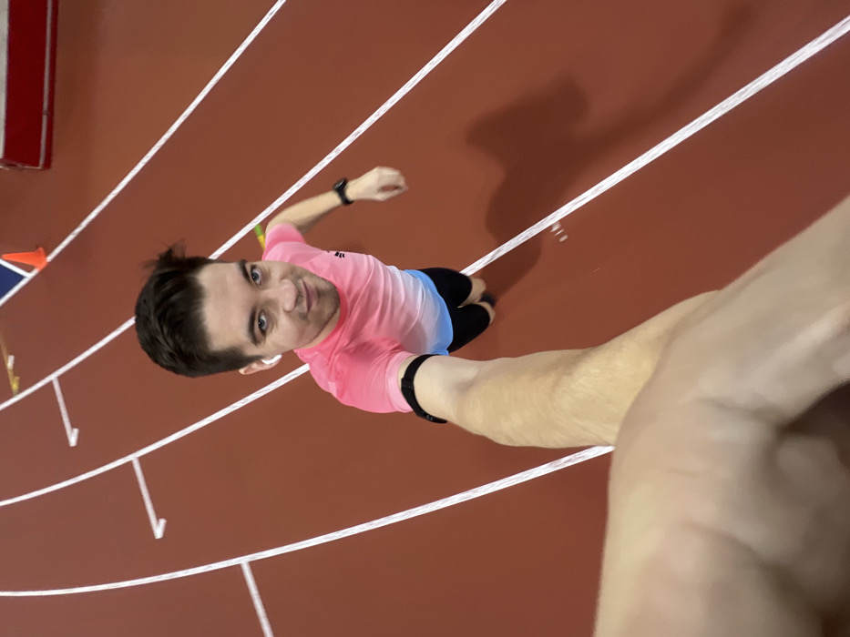 A high-angle selfie captures a person standing on an indoor running track. The camera is held by the person's outstretched right arm, pointing downwards, creating a slightly distorted perspective where the person's head appears larger and their body tapers towards their feet.
The person is a male with dark hair, looking up at the camera with a slight smile. He is wearing a short-sleeved athletic t-shirt that transitions from a vibrant pink at the top to a light blue at the bottom. He has black shorts on and black athletic shoes. On his left wrist, he wears a black fitness tracker or watch, and a white earbud is visible in his left ear. Another black wristband is on his right wrist, which is holding the camera.
The setting is a reddish-brown synthetic running track, marked with crisp white lane lines. The lines are visible across the frame, indicating multiple lanes. The track appears to be indoors, suggested by the uniform lighting and what looks like a wall or barrier on the far left edge of the frame, near an orange cone. The person's shadow is cast prominently on the track behind and to their right, elongated due to the overhead light source. No text is visible in the image. The overall impression is one of a brief pause during an athletic activity, possibly before or after a run or workout.