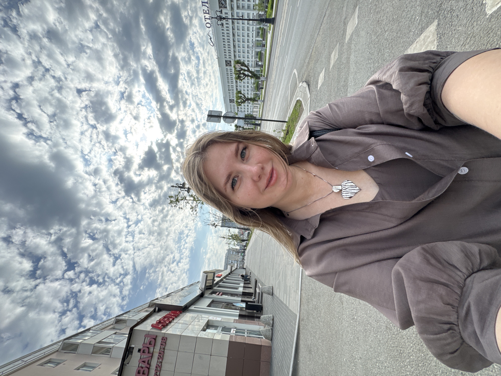 A medium, close-up selfie shot shows a light-skinned woman with blonde hair. She is wearing a collared, long-sleeved shirt and a silver necklace. In the background, a city scene unfolds under a cloudy sky. The "ОТЕЛЬ" hotel can be seen in the distance, alongside a building with the Russian words "ВАРЫ". The scene takes place in the city of Perm, Russia.