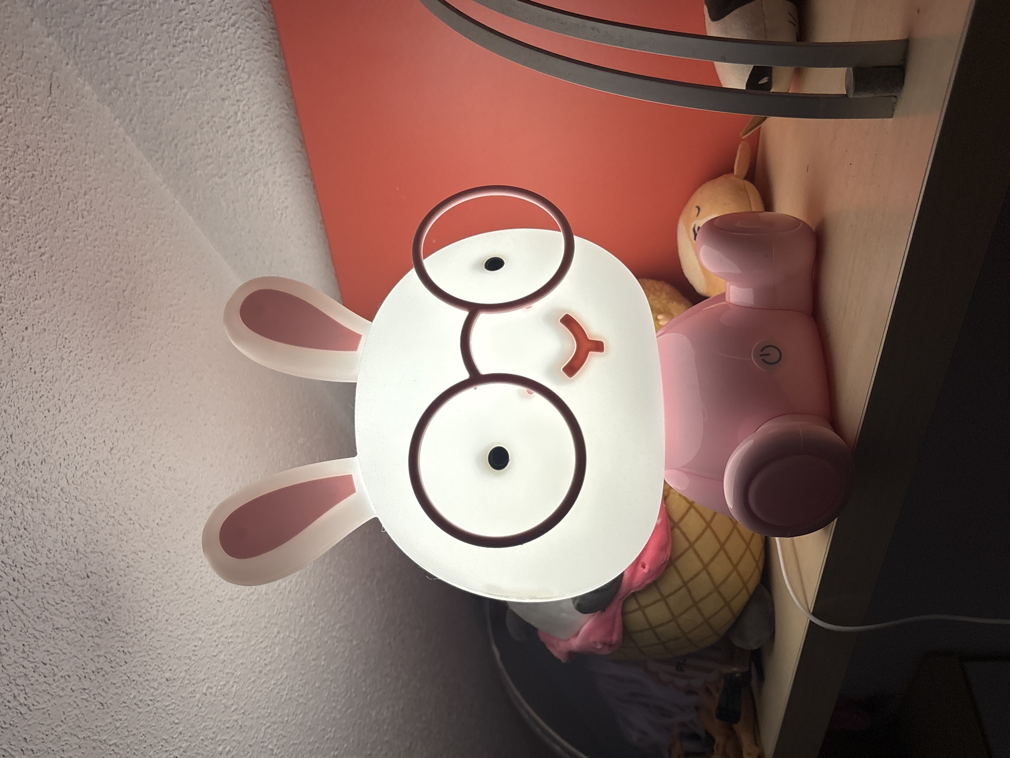 A close-up, low-angle shot captures a white bunny-shaped lamp with pink inner ears and a pink body, sitting on a light wood shelf. The lamp features large, round, dark-rimmed glasses over its eyes, a small orange mouth, and a power button on its belly. To the left of the lamp, partially obscured, is a stuffed toy resembling an ice cream cone with a pink top and yellow cone. Behind the lamp, a red wall is visible. To the right of the lamp, out of focus, are other stuffed toys, including one with a black and white face, and part of a gray metal lamp stand. The shelf appears to be in a dimly lit room with a textured ceiling above. The image location is not specified in the description.