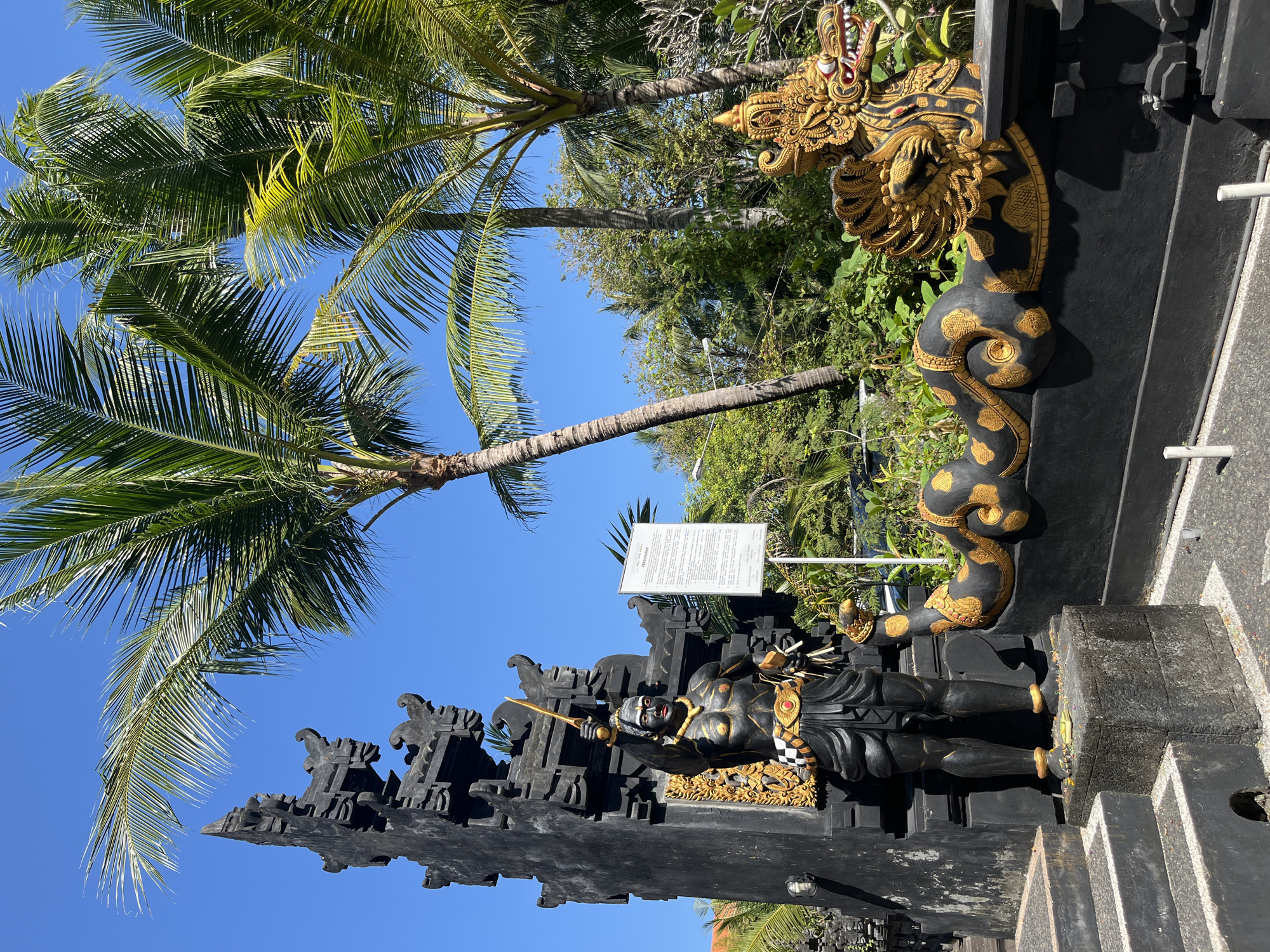 In Kuta Selatan, Indonesia, a dark, ornate statue stands guard in front of lush tropical foliage and towering palm trees under a clear blue sky. The statue depicts a muscular figure with dark skin and red accents on its eyes and lips, wearing traditional Balinese attire. It holds a golden sword aloft in its right hand. To the right of the statue, a decorative black wall features a golden dragon head with intricate details and a coiled serpentine body adorned with golden circular patterns. A white informational sign stands to the right of the statue, providing context about the scene. The ground is paved and scattered with what appear to be colorful flower petals, possibly offerings. Steps lead up to the left of the statue. The overall scene conveys a sense of cultural heritage and tropical beauty.