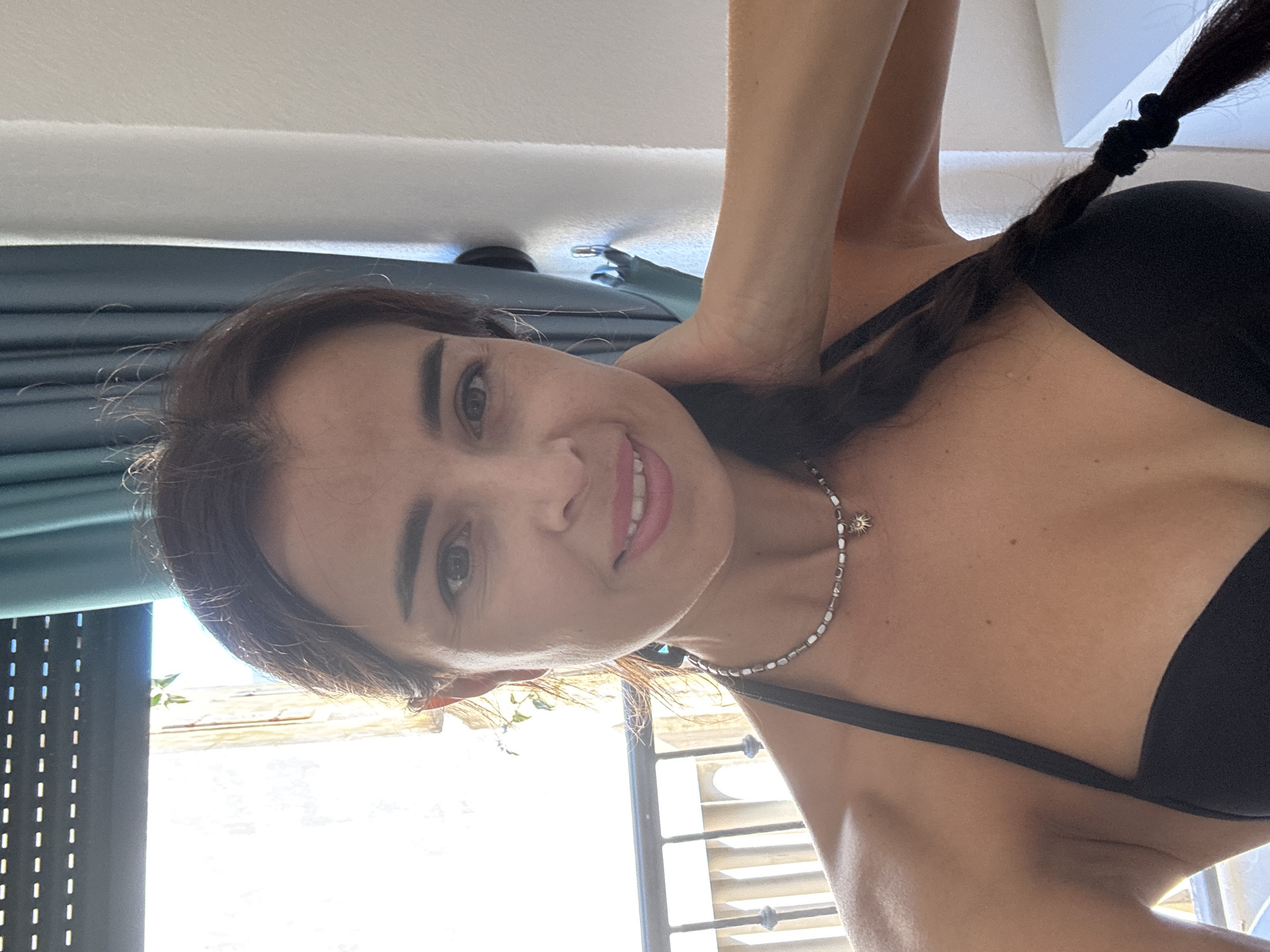 A woman with brown hair, braided and resting over her shoulder, is pictured from the chest up. She is wearing a black bikini top and a beaded necklace with a sun charm. She is smiling and touching her neck with her right hand. Behind her, a window with blinds is visible, along with teal curtains and a balcony railing. The scene appears to be indoors with natural light coming from the window.