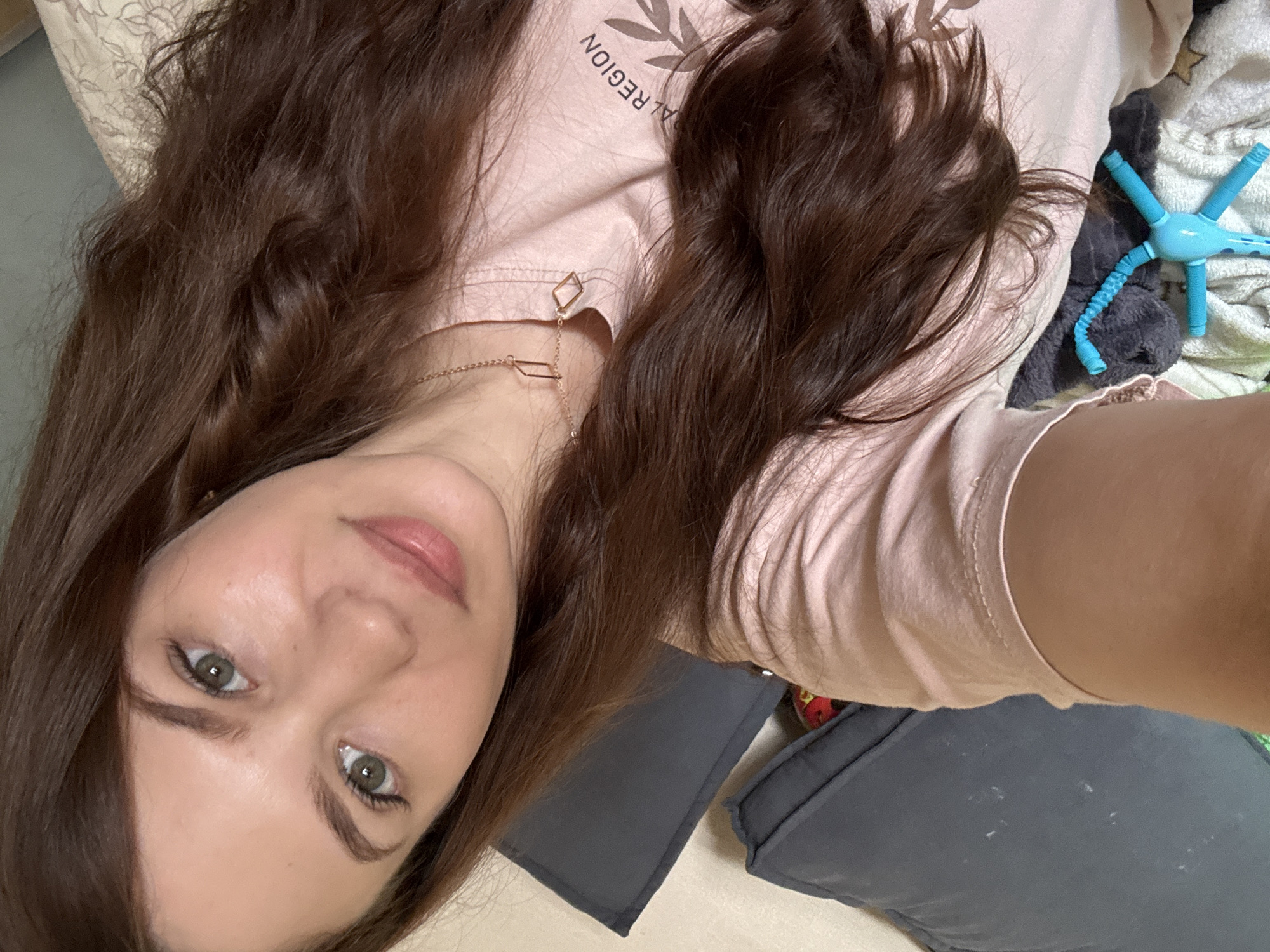 The image is a close-up, overhead selfie of a young woman with long, dark brown wavy hair. She is lying down, with her head tilted upwards and her face angled towards the camera. Her eyes are green and she has light makeup on, including mascara and lip gloss. She is wearing a light pink t-shirt with some text printed on it. A delicate gold necklace with geometric pendants rests on her collarbone.
The background suggests an indoor setting, likely a bedroom. She is positioned on pillows, with one dark gray pillow visible beneath her. To the right of her head, a bright blue hair curler, shaped like a star with radiating tubes, is visible against a soft, textured blanket. The overall lighting appears to be natural, possibly from a window, and the scene conveys a relaxed and casual mood. There is some visible text on her t-shirt, which reads "AL REGION" with a stylized leaf graphic above it. The location is identified as Chaikovsky, Russia.