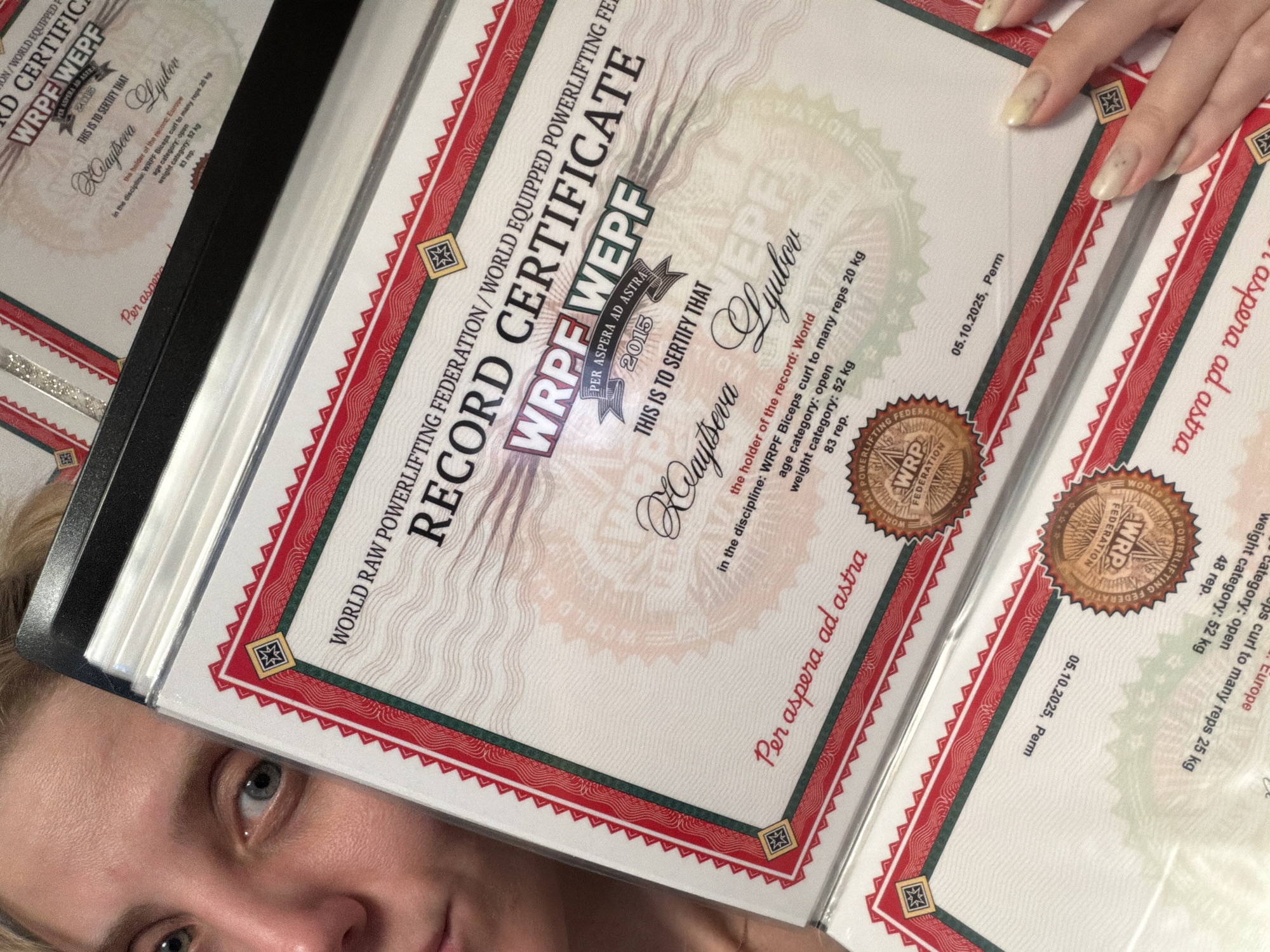 A close-up, angled shot captures a woman partially obscured by several powerlifting record certificates displayed in a holder. The certificates, from the "WORLD RAW POWERLIFTING FEDERATION / WORLD EQUIPPED POWERLIFTING FE WRPF WEPF," are the central focus, celebrating significant athletic achievements.
The most prominent certificate is a "RECORD CERTIFICATE" that identifies "Waytseva Lyubov" as the "World" record holder. It specifies her accomplishment in the "WRPF Biceps curl to many reps 20 kg" discipline, within the "open" age category, weighing "52 kg," with an impressive "83 reps." This certificate also bears the motto "PER ASPERA AD ASTRA" and the year "2015." Another visible certificate confirms a "Europe" record for the same individual, discipline, and stats, dated "05.10.2025, Perm."
The woman's face is visible in the bottom left, with one clear blue/gray eye looking towards the viewer, conveying a sense of pride or quiet accomplishment. Her light-colored hair and the tips of her fingers, adorned with light-colored nail polish, are also seen holding the certificates in the top right. The setting appears to be indoors. The certificates list "Perm" as a location, which is a city in Russia, aligning with the provided location context of Chaikovsky, Russia.