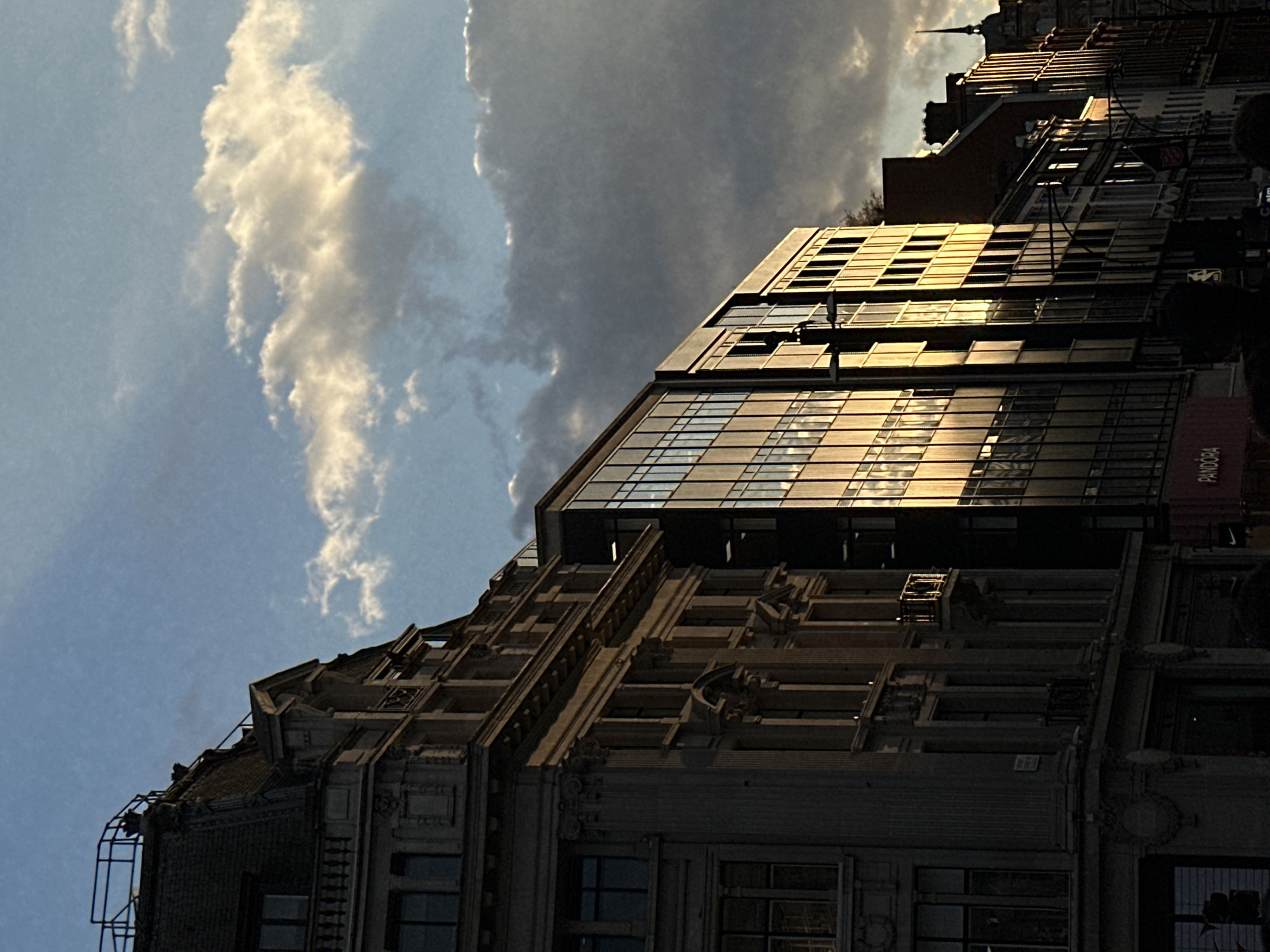 The image depicts a building with a modern glass extension atop an older, ornately decorated structure. The glass facade reflects sunlight. A sign reading "PANDORA" is visible on the building. The sky shows both dark, cloudy areas and patches of blue with white clouds. The location is London, United Kingdom, specifically a commercial area with other buildings visible in the background. Pedestrians can be seen on the right-hand side of the frame.